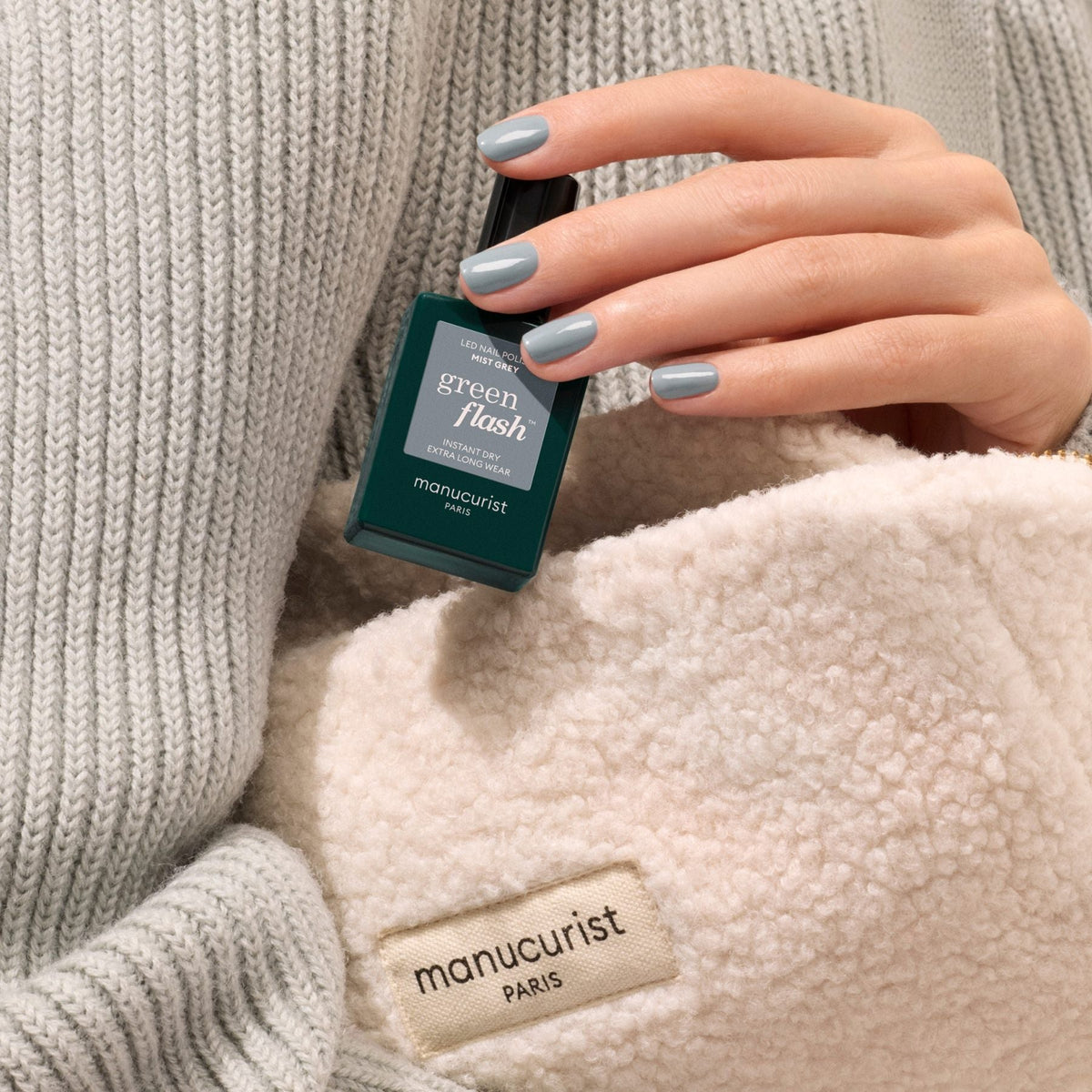 A hand with Mist Grey gel polish by Manucurist holds a matching bottle. The person wears a gray knit sweater and a cream fleece featuring the Manucurist label.