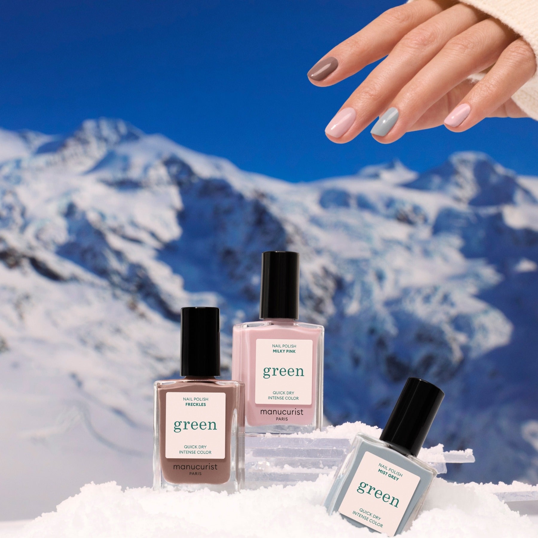 A hand with neatly painted, neutral nails hovers above Manucurist’s Trio Collections nail polishes set in the snow, reflecting nail trends with snowy mountains and a clear blue sky in the background.