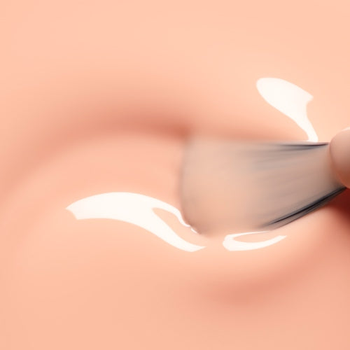 A close-up of a makeup brush applying a glossy, creamy substance onto smooth, peach-colored skin evokes the soft radiance of Manucurist’s Milky Peach gel polish.