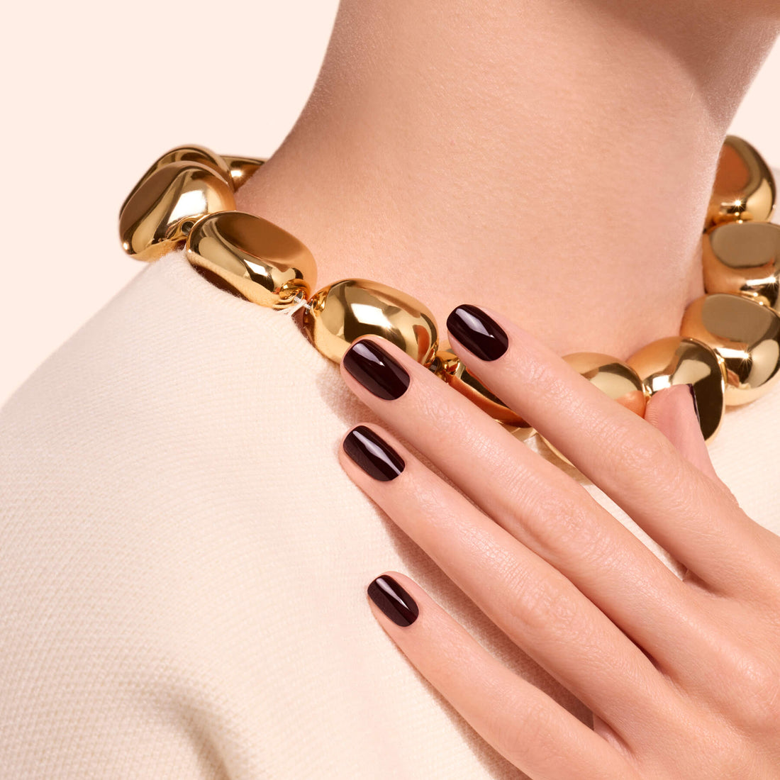 A woman wearing a chunky gold necklace and a cream top displays her hand with nails painted in Hollyhock by Manucurist, featuring a deep red polish, resting near her collarbone.