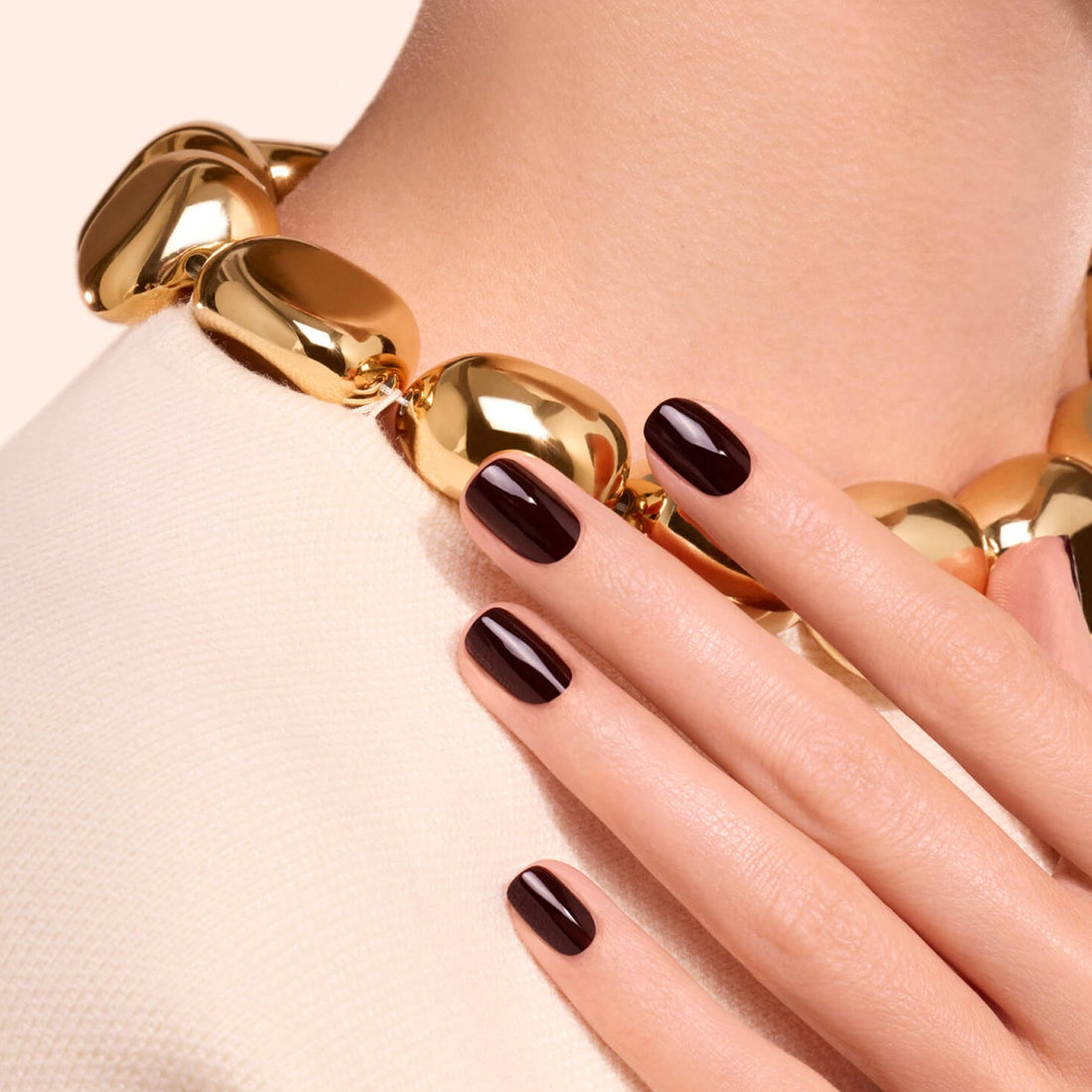 A woman wearing a chunky gold necklace and a cream top displays her hand with nails painted in Hollyhock by Manucurist, featuring a deep red polish, resting near her collarbone.