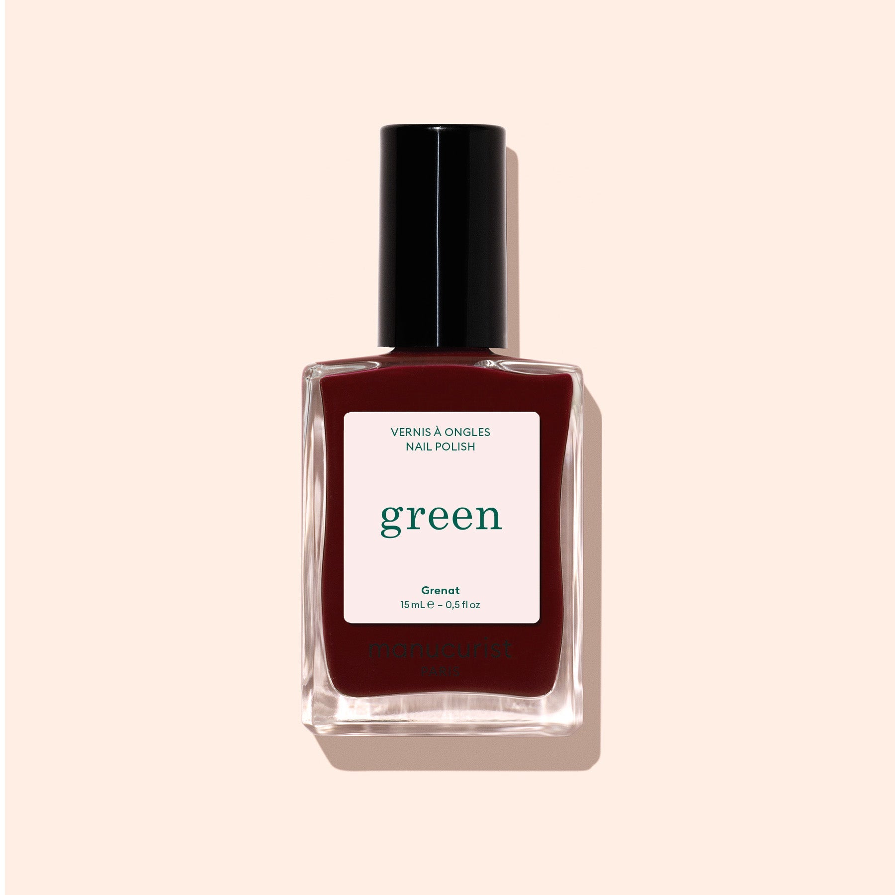 A bottle of Manucurist Grenat red nail polish with a black cap, labeled green and Green™, sits on a light beige background.