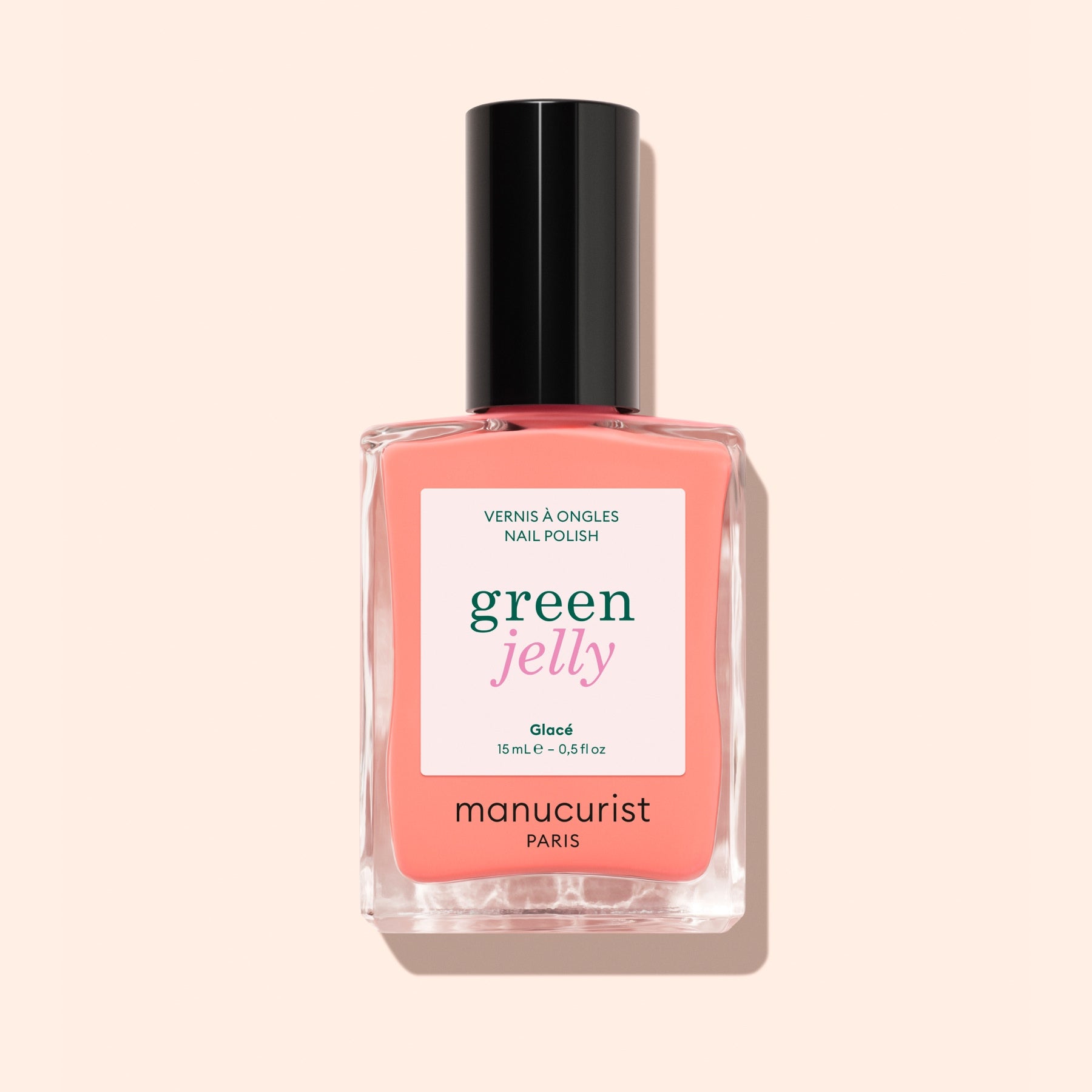 A bottle of Manucurist Glacé ultra-glossy jelly nail polish in coral pink with a black cap, shown against a light peach background.