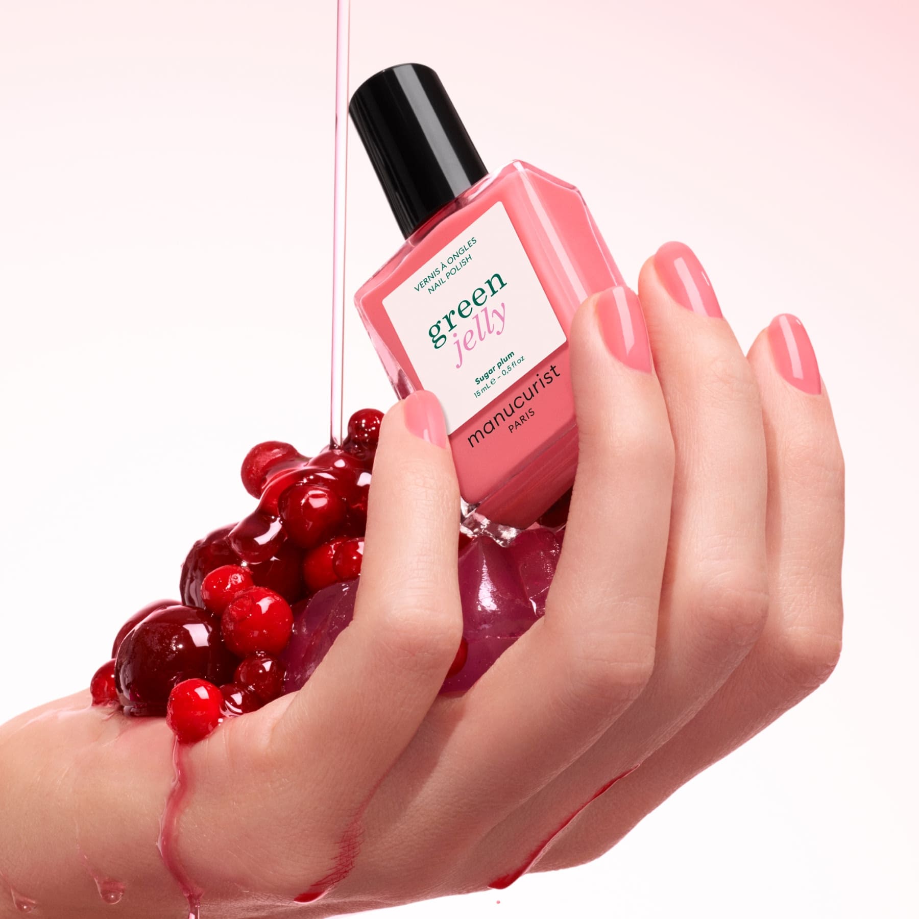 A hand with neatly manicured pink nails holds shiny red berries and a bottle of Manucurist’s Sugar Plum nail polish, as pink liquid drips for an ultra-glossy finish against a soft pink background.