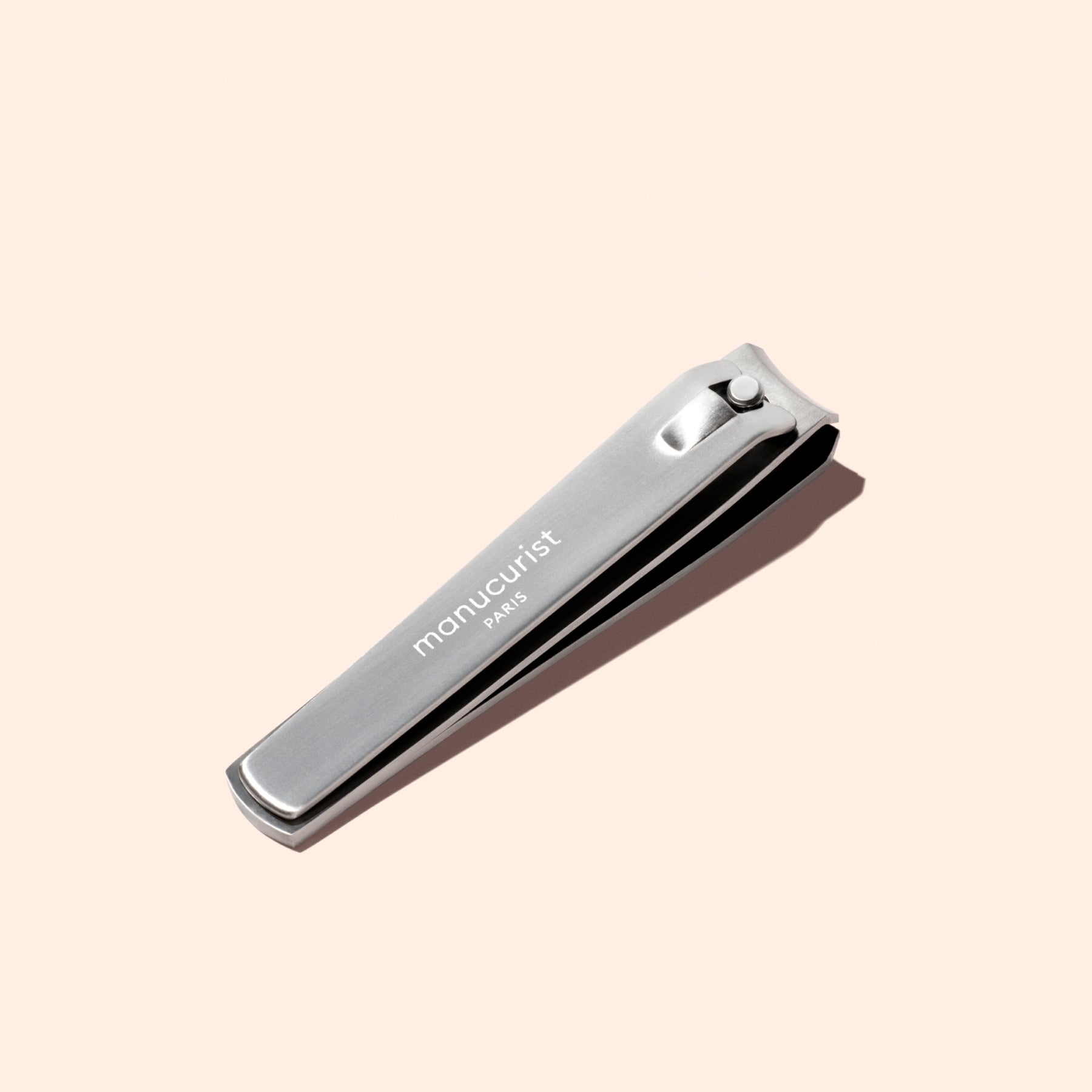 A silver Manucurist Nail Clipper, designed for precise manicures and ideal for shaping natural nails, is engraved with the brand name and displayed on a light beige background.