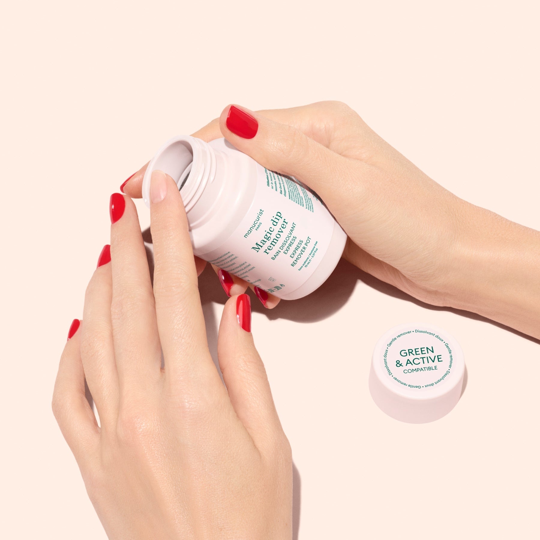 A person with red nails holds a travel-friendly, acetone-free Manucurist Magic Dip Remover in a white bottle with the lid off, set nearby on a light beige background.