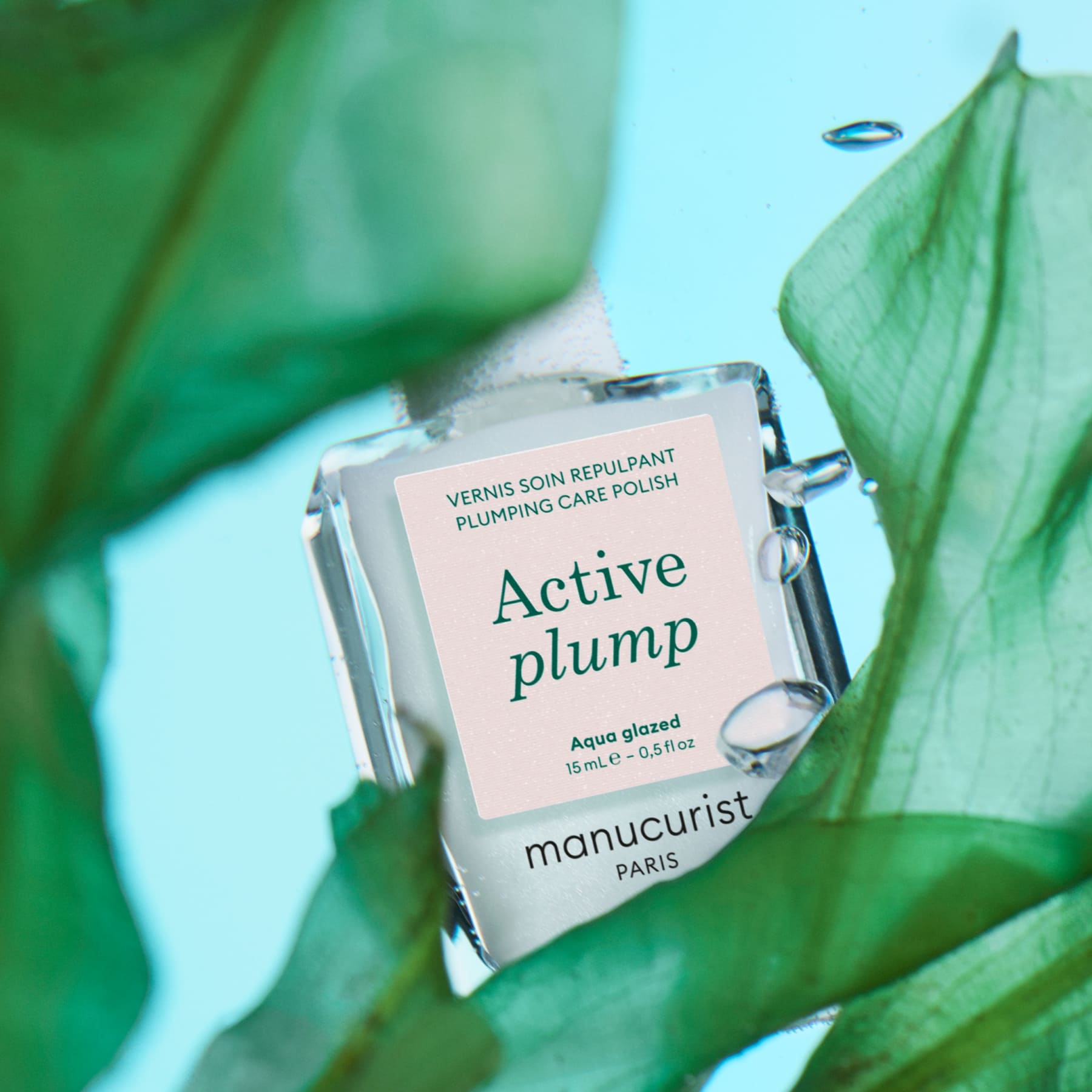 Manucurist Active Plump Aqua Glazed nail polish is shown among green leaves with water droplets on both, set against a light blue background, highlighting the formula’s hydrating benefits for nails.