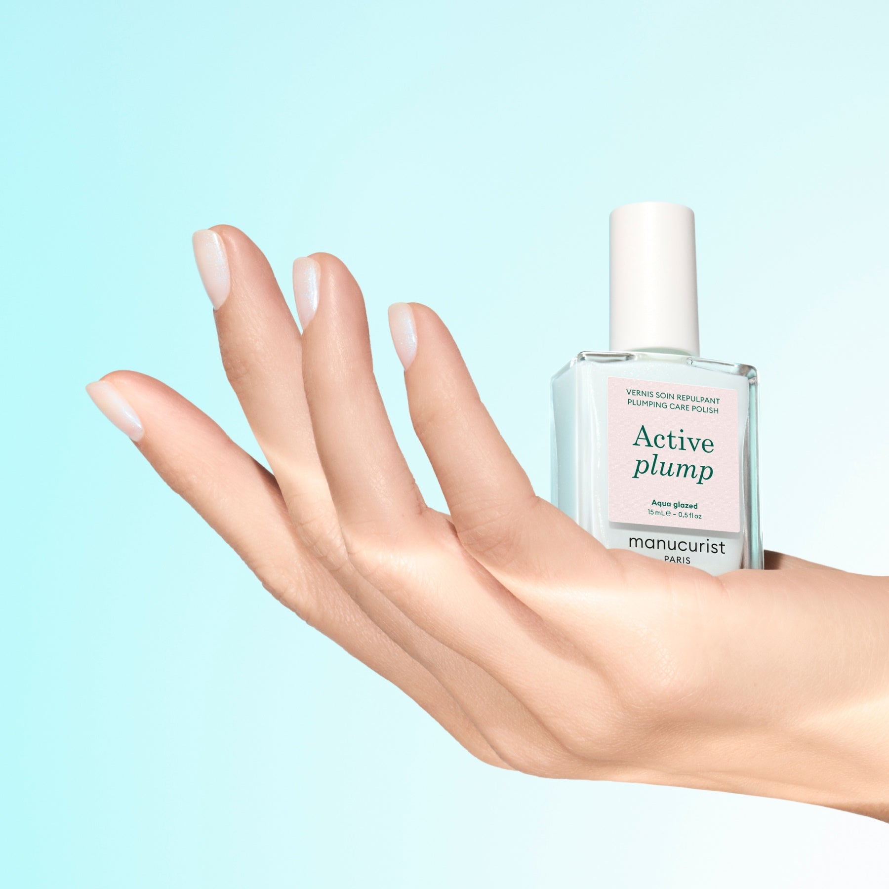 A hand with hydrated, pale pink nails holds a bottle of Manucurist Active Plump Aqua Glazed nail polish against a light blue gradient background.