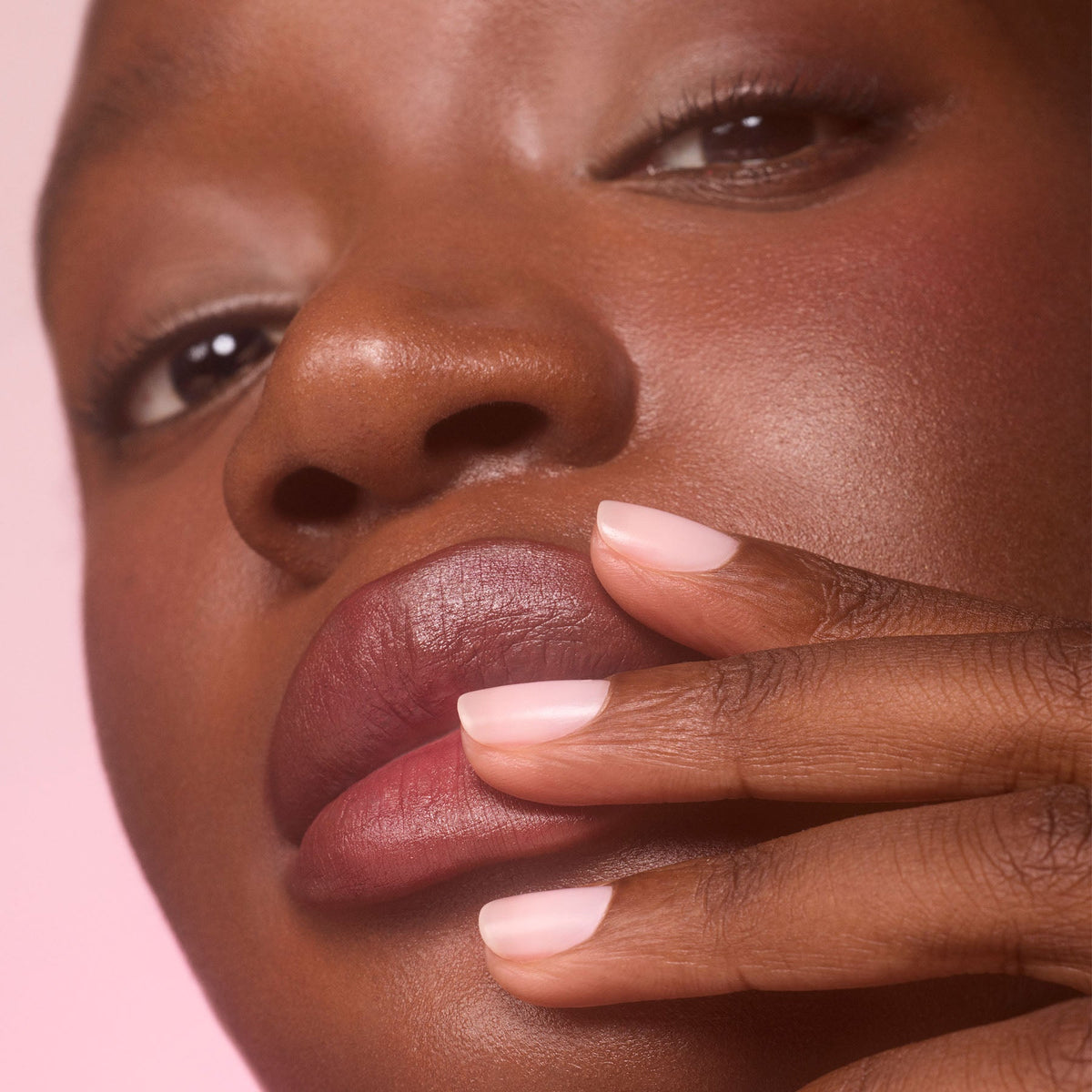 A close-up of a person with dark skin touching their lips with manicured fingers, their short nails polished with Manucurist’s Active™ Blur matte nail polish. The softly blurred background features light pink tones.
