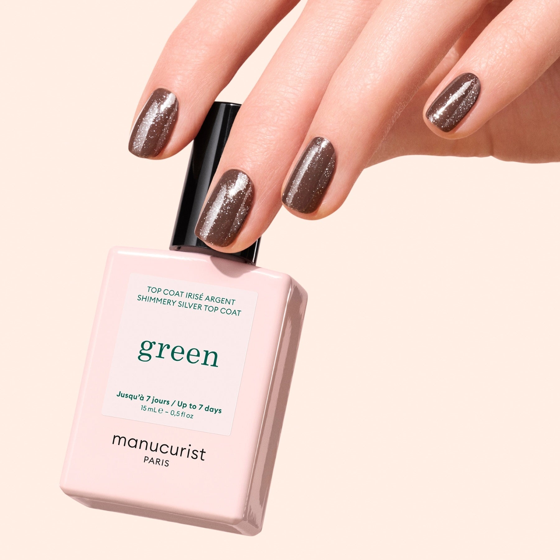 A hand with short, neatly manicured nails painted in shimmery brown holds a bottle of Manucurist Glitter Top Coat against a soft beige background. The gel-like finish adds subtle green glitter for extra shine.