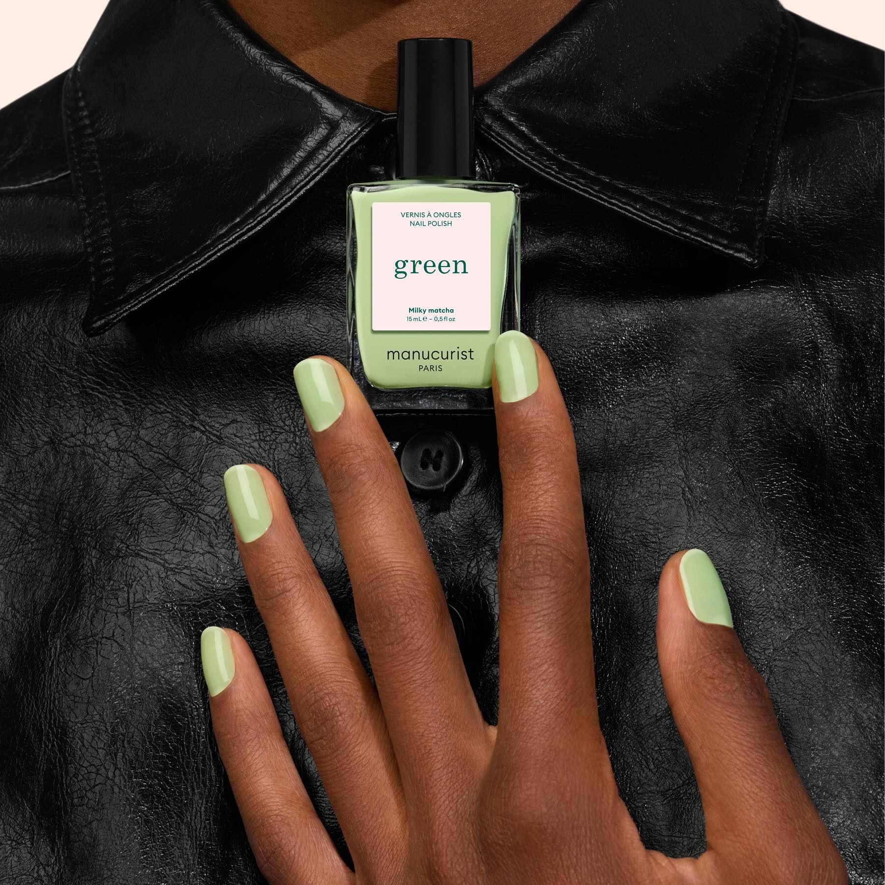 A hand with light green polished nails holds a bottle of Manucurist’s Milky Matcha nail polish, perfectly matching the manicure, set against a black leather shirt background.