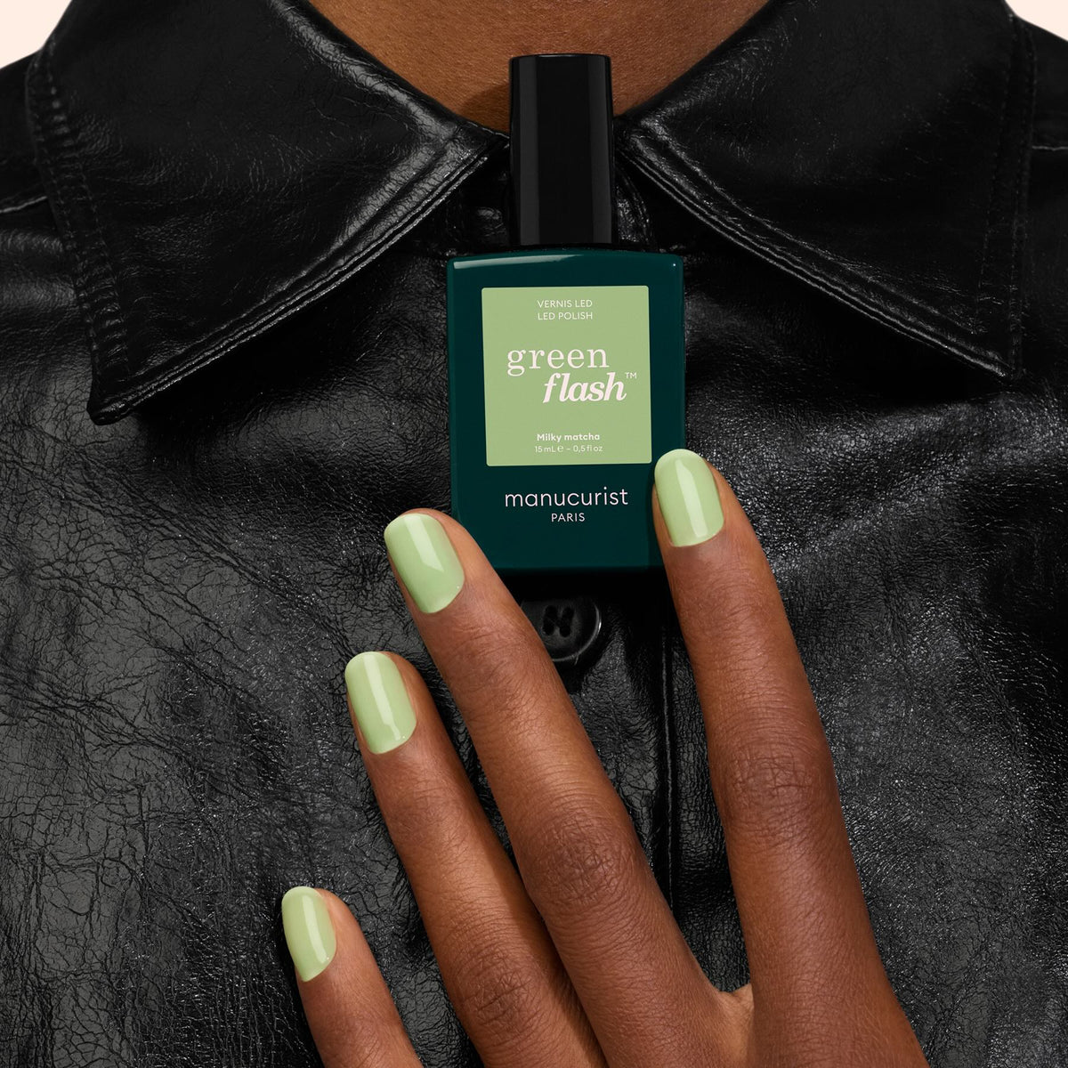 A hand with pastel green painted nails holds a bottle of Milky Matcha nail polish by Manucurist, featuring creamy nuances and subtle botanical notes, against a black leather jacket.