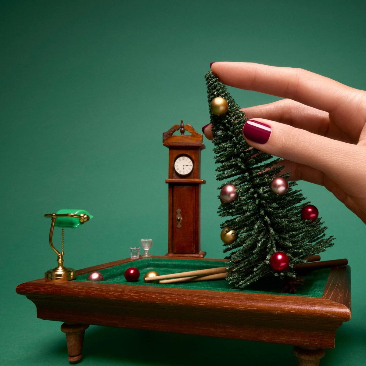 A hand with luminous burgundy nails, polished to perfection with Manucurist’s Amour Green Flash gel polish, places a small decorated Christmas tree on a miniature billiards table beside a tiny lamp, two glasses, and a grandfather clock, all set against a green background.