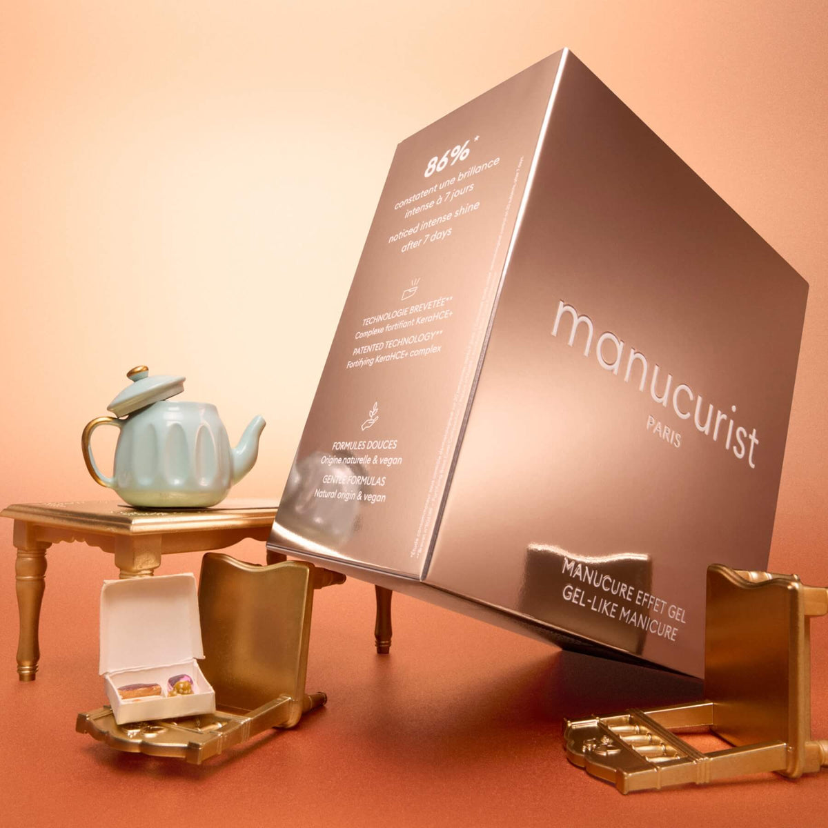 A large metallic pink box labeled “Green™ Gift Set Holiday” by Manucurist sits among gold vanity items and a blue teapot on a peach-colored surface with an orange gradient background.