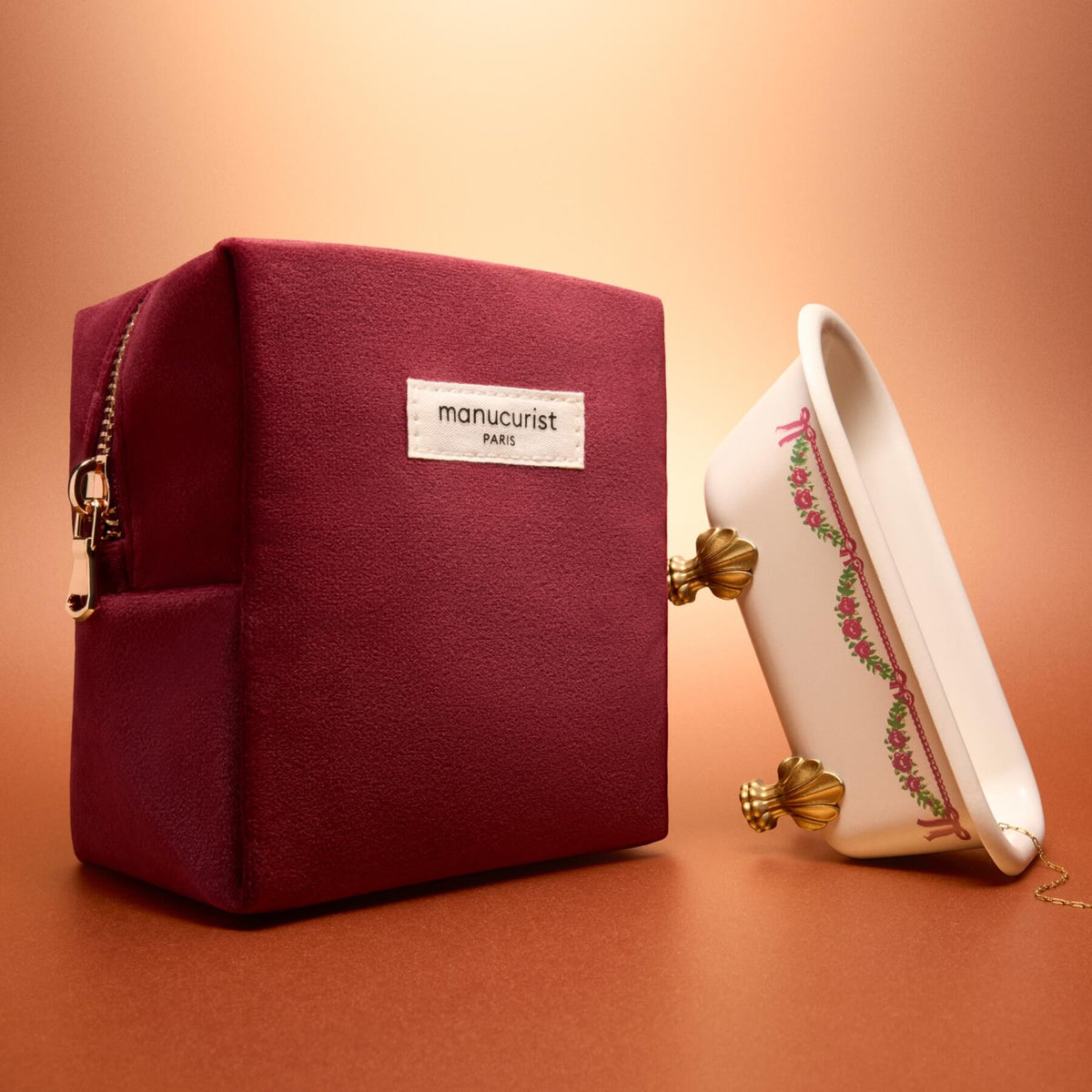 A Manucurist Velvet Pouch Holiday rests next to a small decorative porcelain bathtub with gold claw feet, set against a warm brown background.