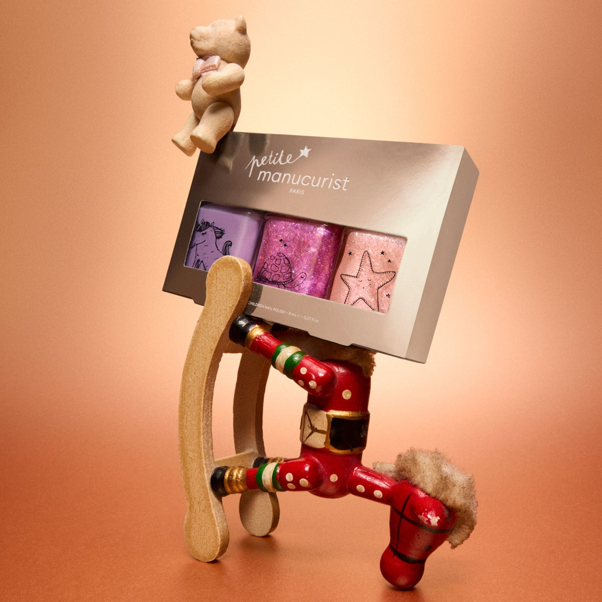 A whimsical scene featuring a gingerbread sled, the legs of a red wooden nutcracker, and a bear holding the Petite Manucurist Trio by Manucurist—three pastel children’s nail polishes made from natural origin ingredients—set against a warm orange background makes this set an ideal holiday gift for little ones.