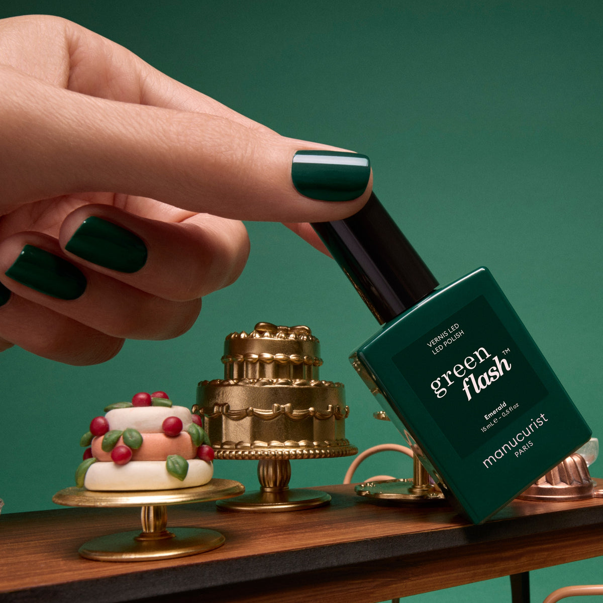 A hand with deep forest green Emerald by Manucurist manicured nails holds a bottle of Green Flash LED gel nail polish above miniature decorative cakes on a small wooden tray.