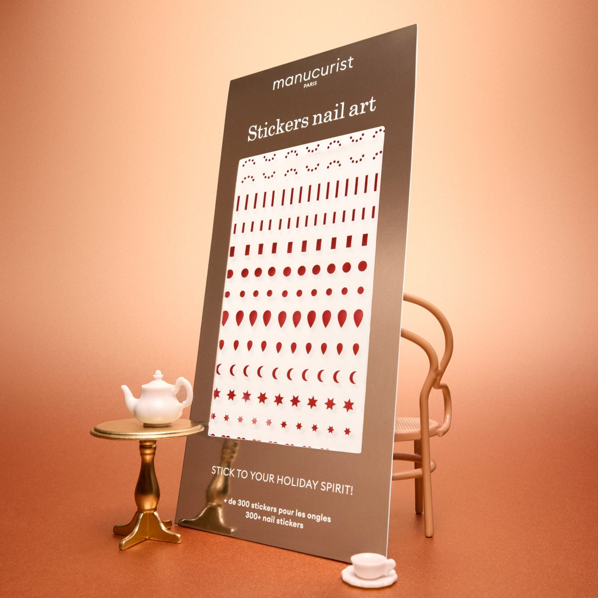 A large card featuring Manucurist Stickers Nail Art Holidays is displayed in a warm, peach-colored setting, propped on a table with a small teapot, cup, and nearby chair. The card showcases festive red nail art stickers in various shapes—perfect for creating celebratory manicures. Text on the card reads “manucurist Stickers nail art.”.