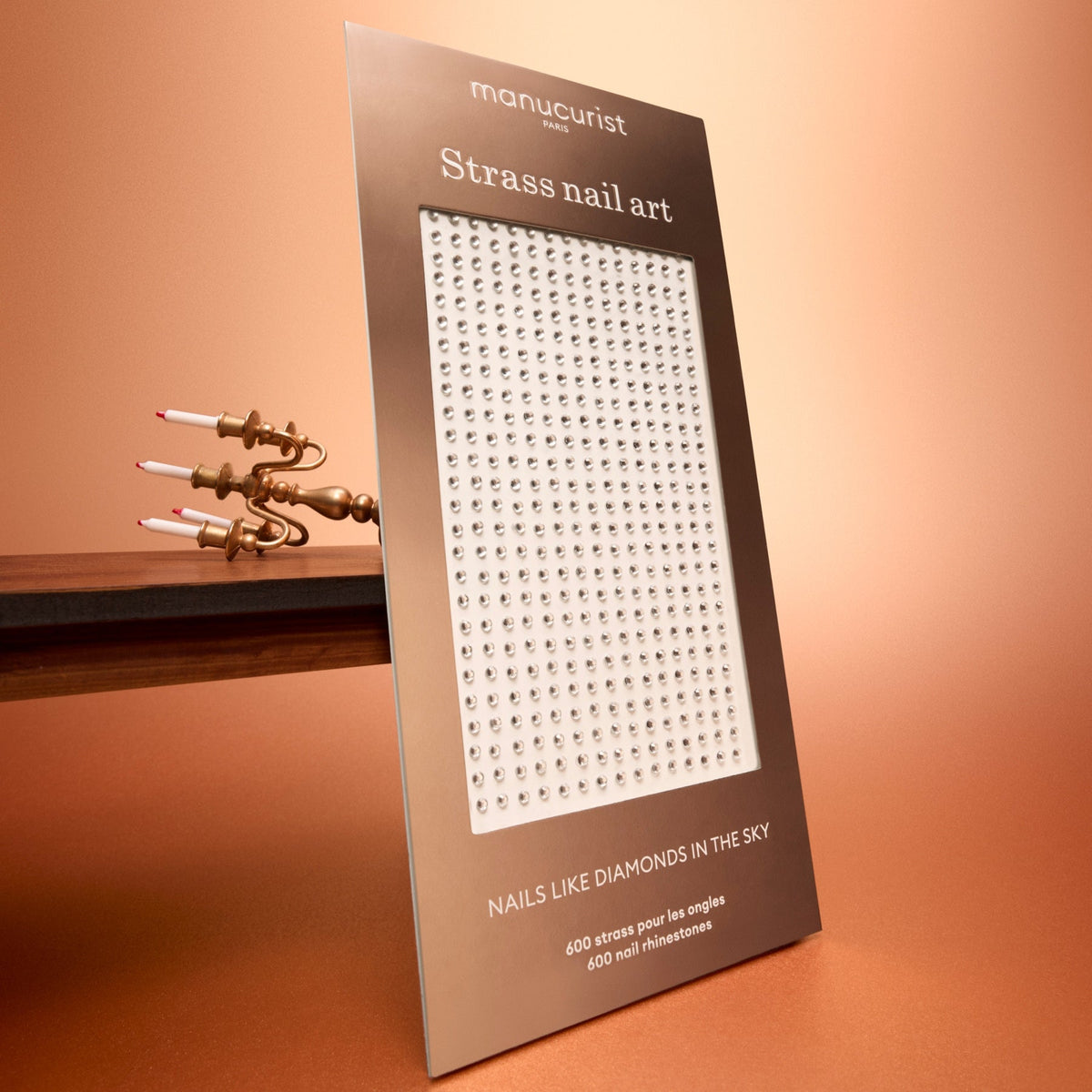 A package of Manucurist Rhinestones Nail Art Holidays is displayed upright on a brown surface, with a candleholder in the background against a peach gradient backdrop—perfect for adding festive flair to your holiday manicure.