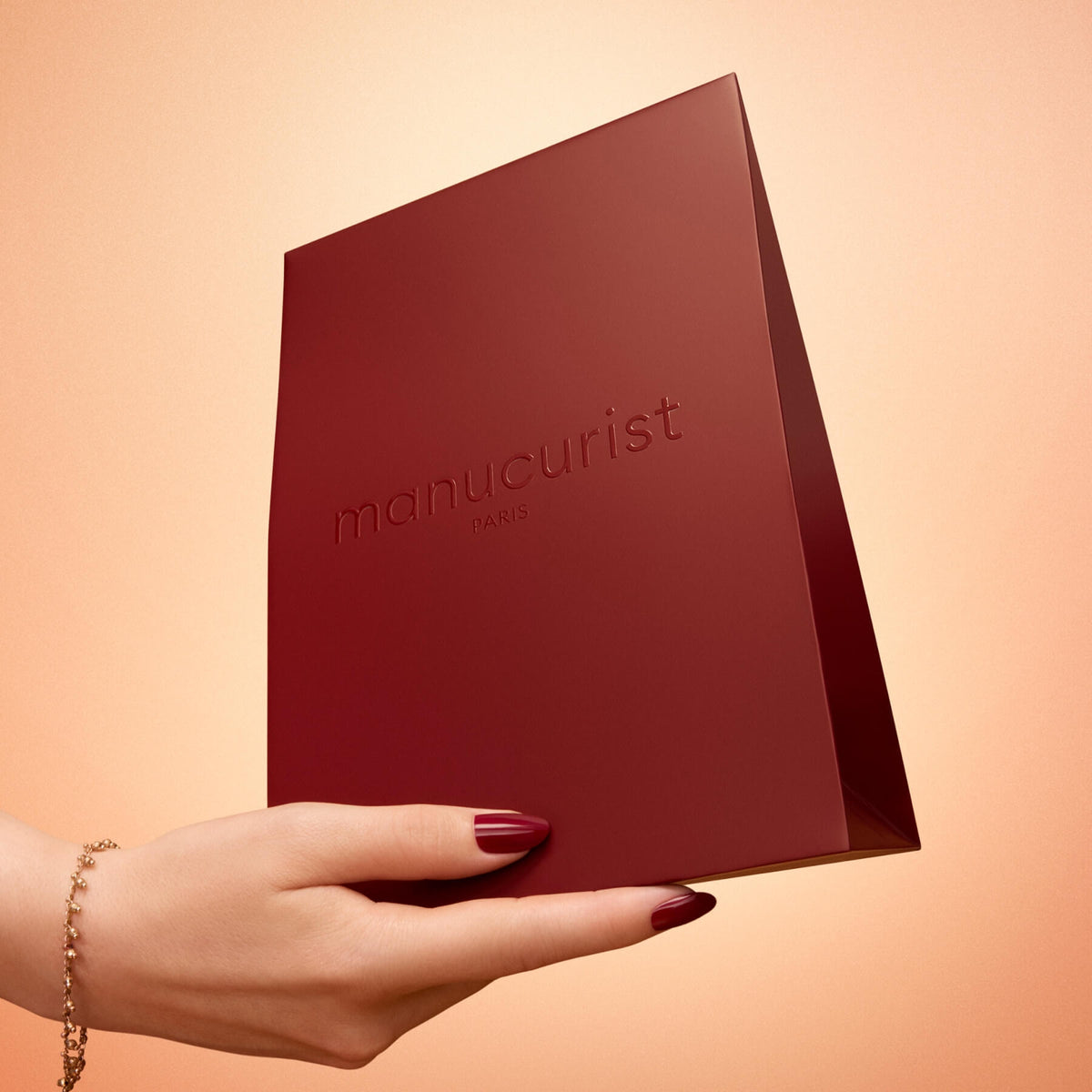 A hand with polished nails and a gold bracelet holds the Holiday Gift Bag - S by Manucurist, ideal for gifting accessories, against a light peach background.