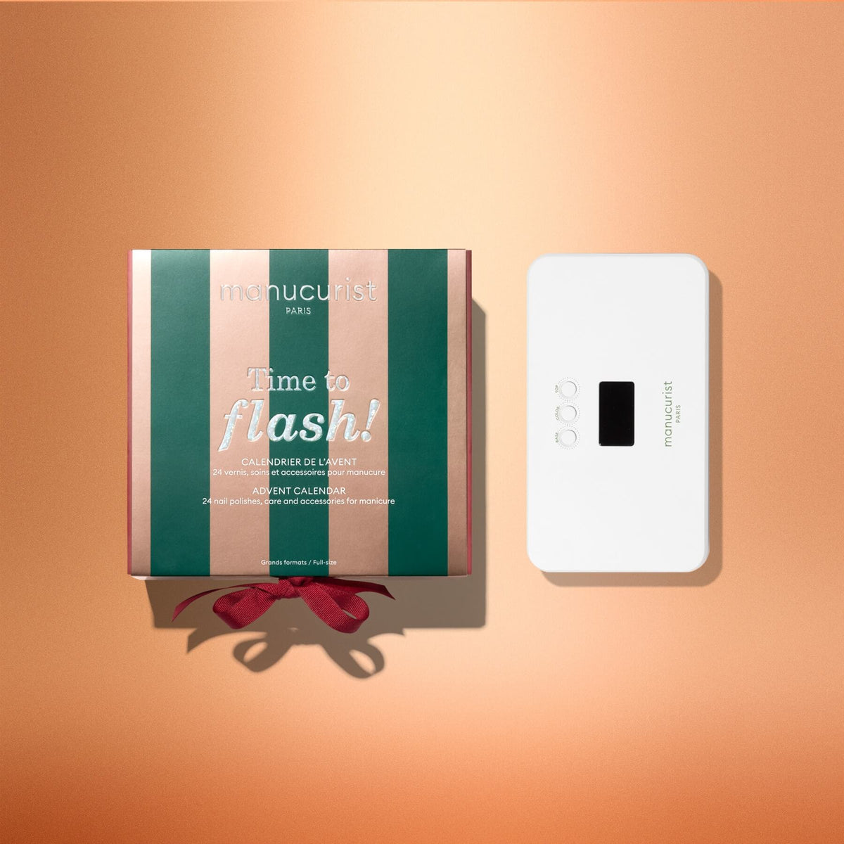 A green and pink striped Manucurist Advent Calendar Time to Flash! box sits next to a small white LED lamp with buttons and a screen, both displayed on a peach-colored background—perfect for creating your next gel polish manicure.