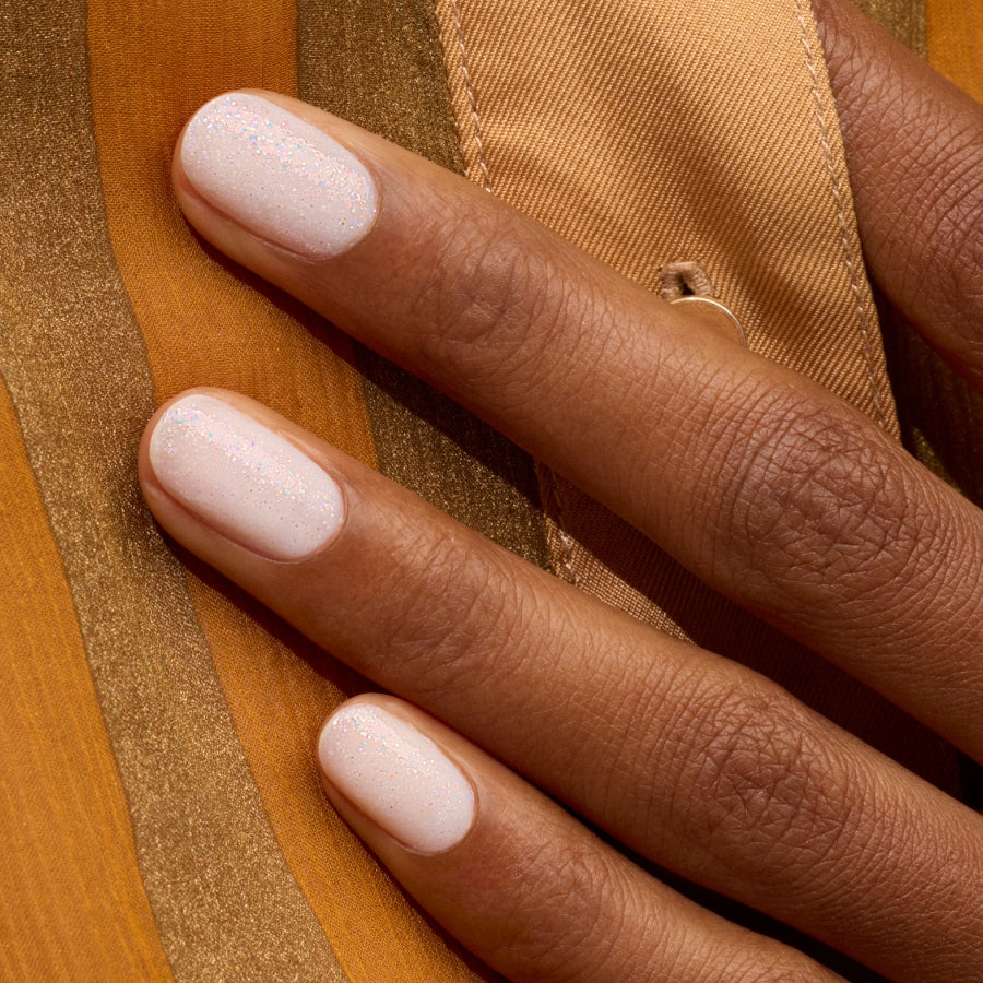 A close-up of a hand with neatly manicured nails painted in Manucurist's Nebula, a soft, shimmery milky white polish, resting on gold and brown striped fabric.