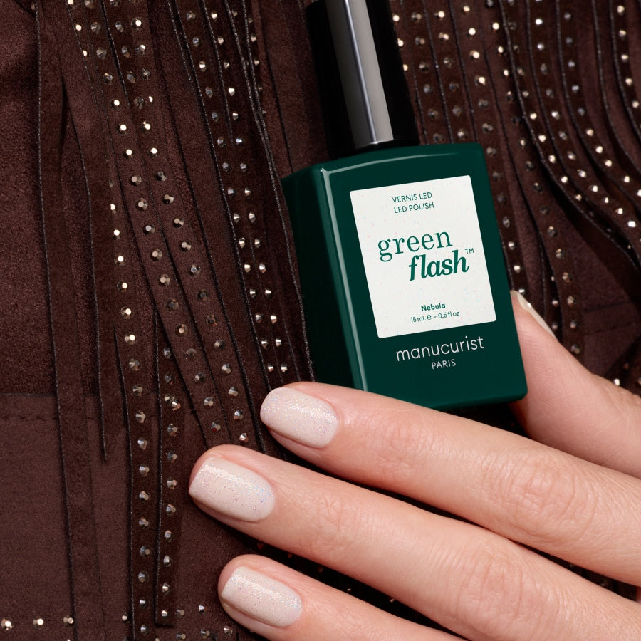 A hand with milky white, shimmery nail polish holds a dark green bottle of Manucurist Nebula nail polish against a brown, fringed, studded fabric background.