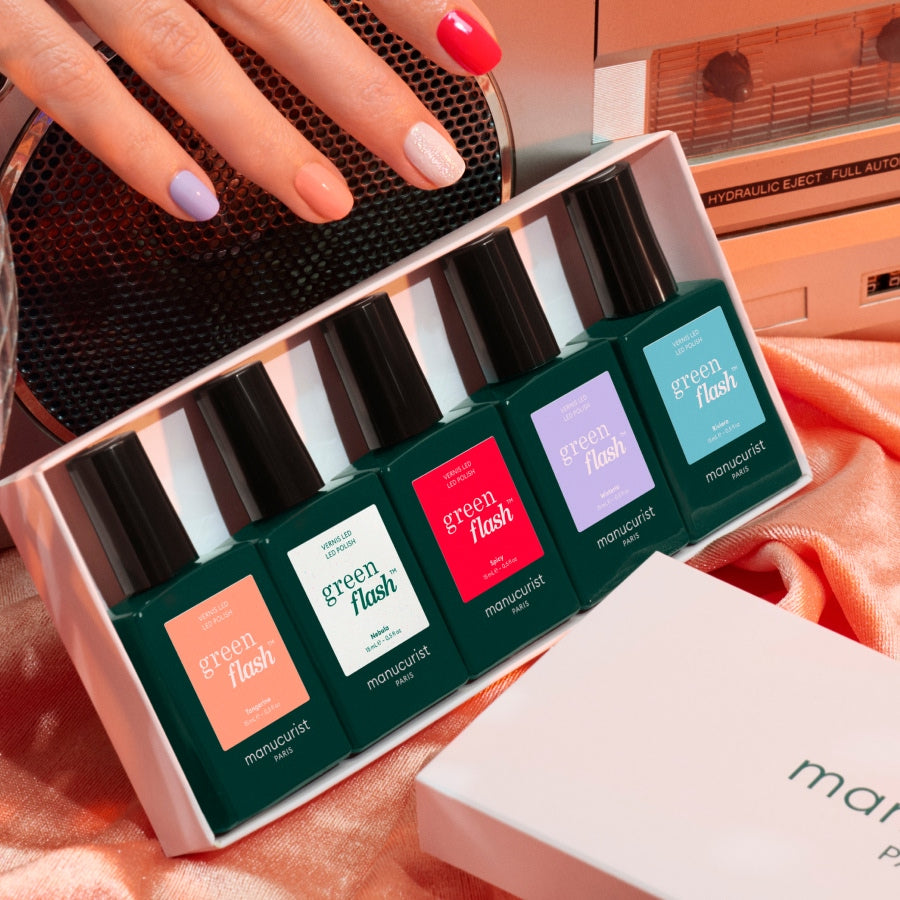 A hand with pastel nails rests near a box of Manucurist Rainbow Collections, featuring five vibrant Green Flash LED nail polishes on pink satin beside a retro stereo, sparking creative manicure color ideas.