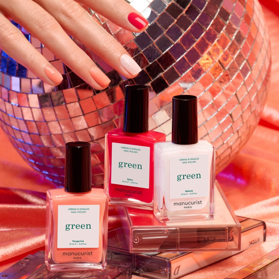 A hand with a pink and white manicure touches a disco ball, while three Manucurist Trio Collections nail polish bottles rest on cassette tapes against a silky coral backdrop, highlighting the latest nail trends.