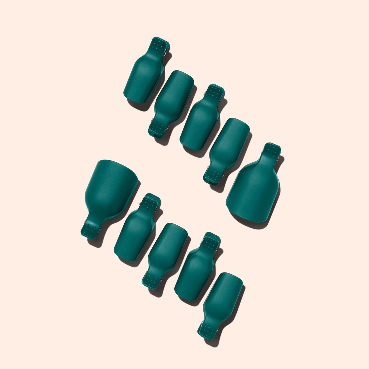 Nine Manucurist Foot Removal Clips, in dark green silicone, are arranged diagonally on a light beige background, casting shadows to the upper left.