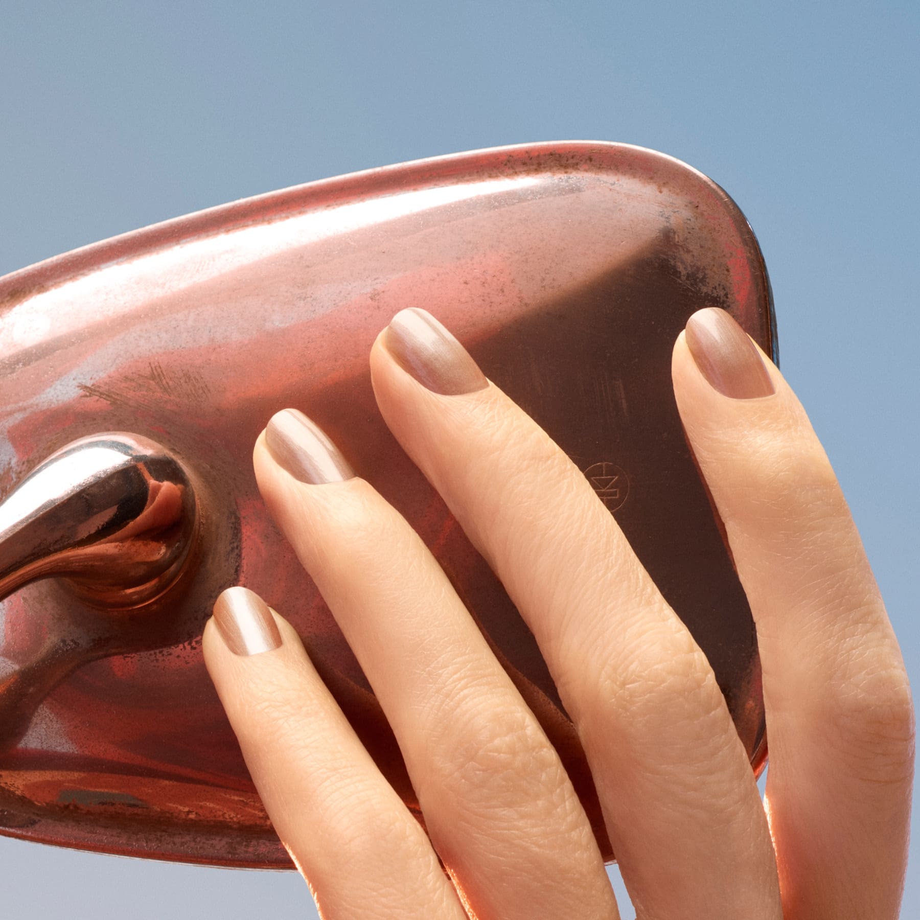 A hand with neatly manicured nails painted in Manucurist’s Sandstone gently touches a shiny, copper-colored object against a clear blue sky.