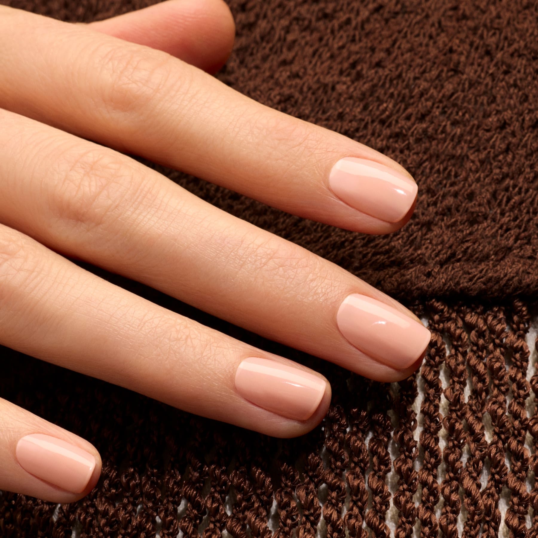 A hand with short, neatly manicured nails painted in Manucurist’s Milky Peach, a glossy orange beige shade, rests on textured brown fabric.