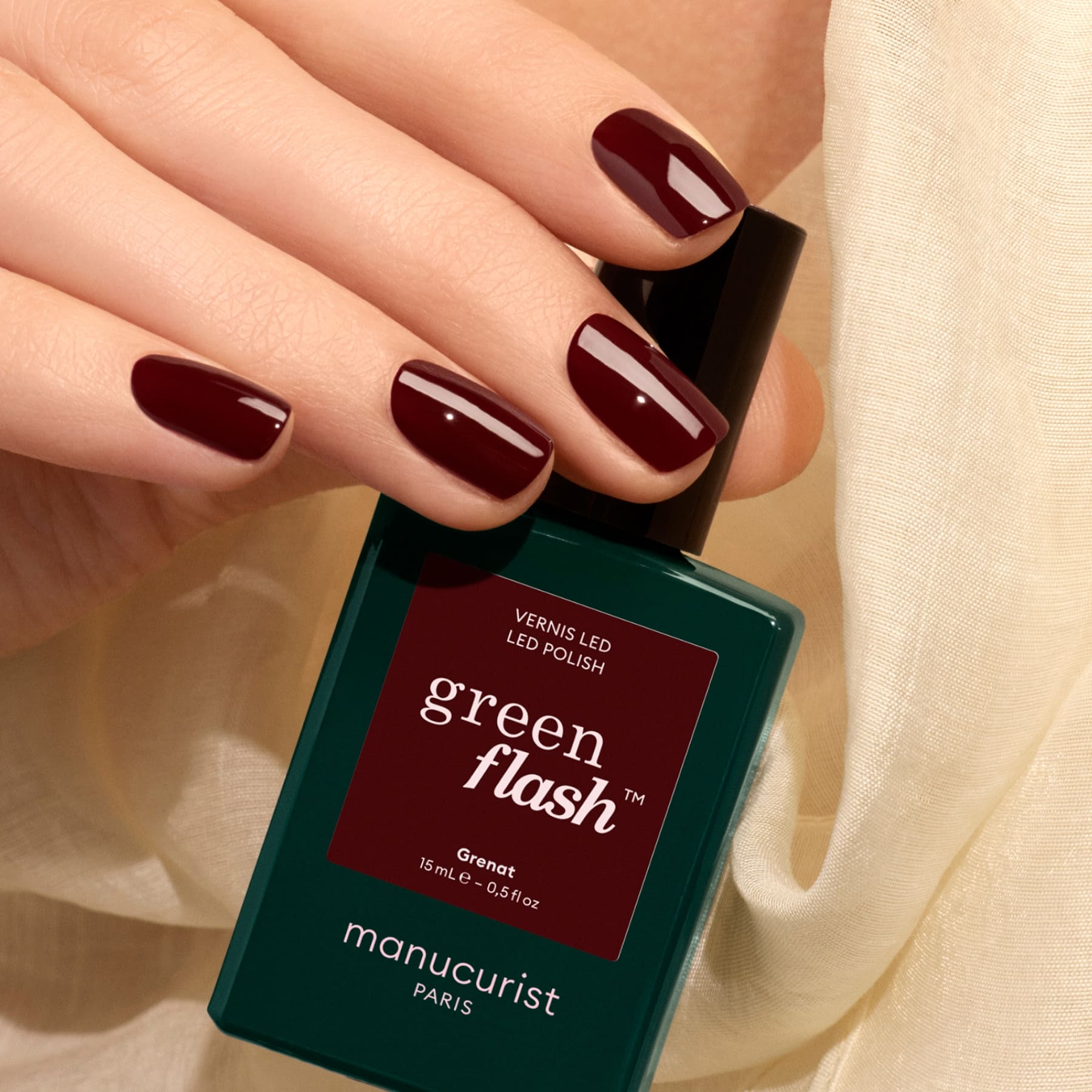 A hand with earthy red nails painted in Manucurist’s Grenat holds a green bottle of gel polish against a light-colored fabric background.