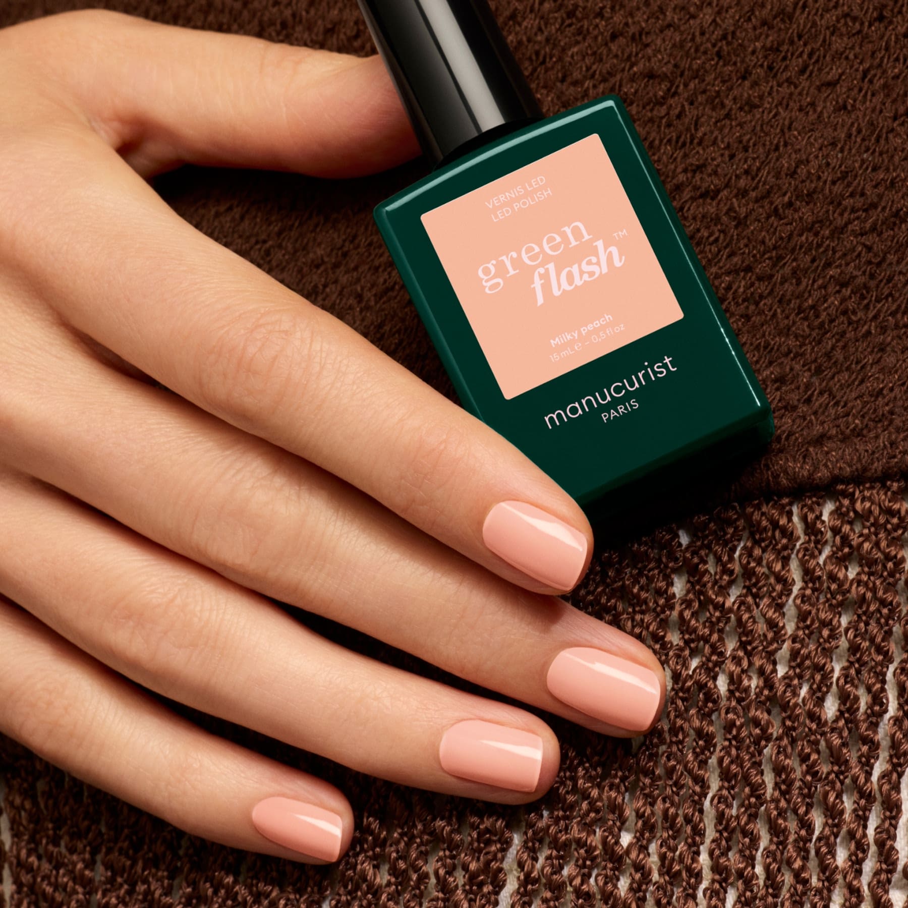 A hand with short, neatly manicured nails painted light pink rests on a brown textured fabric, holding a green bottle of Manucurist nail polish, while shades like Milky Peach by Manucurist and orange beige add to the soft, elegant mood.