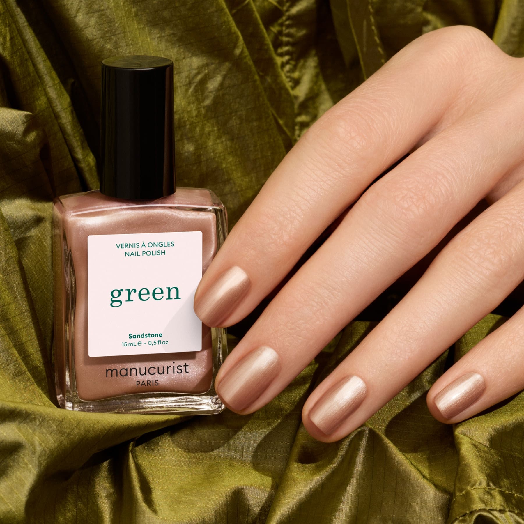 A hand with pearly brown nail polish is displayed next to a bottle of Manucurist Sandstone nail polish, set on an olive green fabric background.