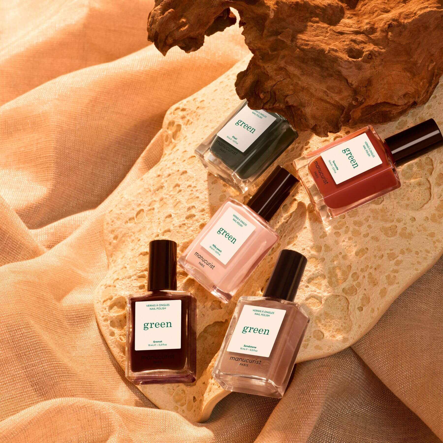 Four bottles of Manucurist’s Rainbow Collections nail polish in earth tones are arranged on a textured stone surface, accompanied by driftwood and set against a beige fabric backdrop, all bathed in warm natural light—an inviting display that highlights this season’s curated colour palettes and the latest trends from Manucurist for effortless manicures.