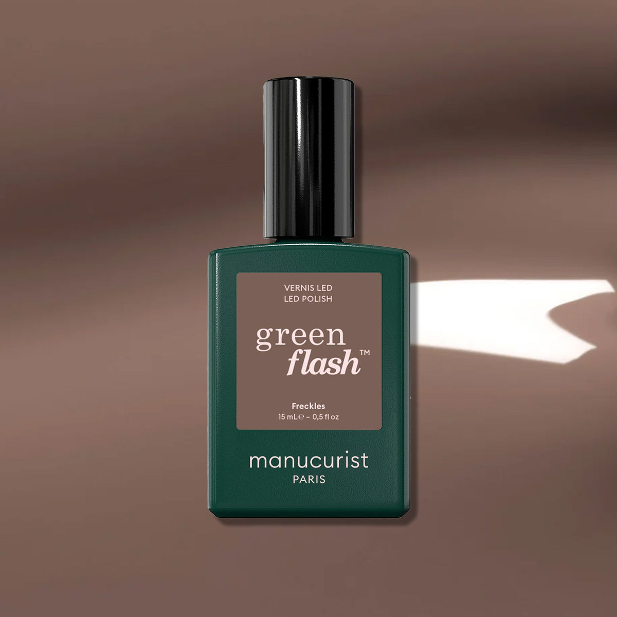 A green Manucurist Freckles nail polish bottle rests on a wood-toned brown surface with a streak of white light beside it.