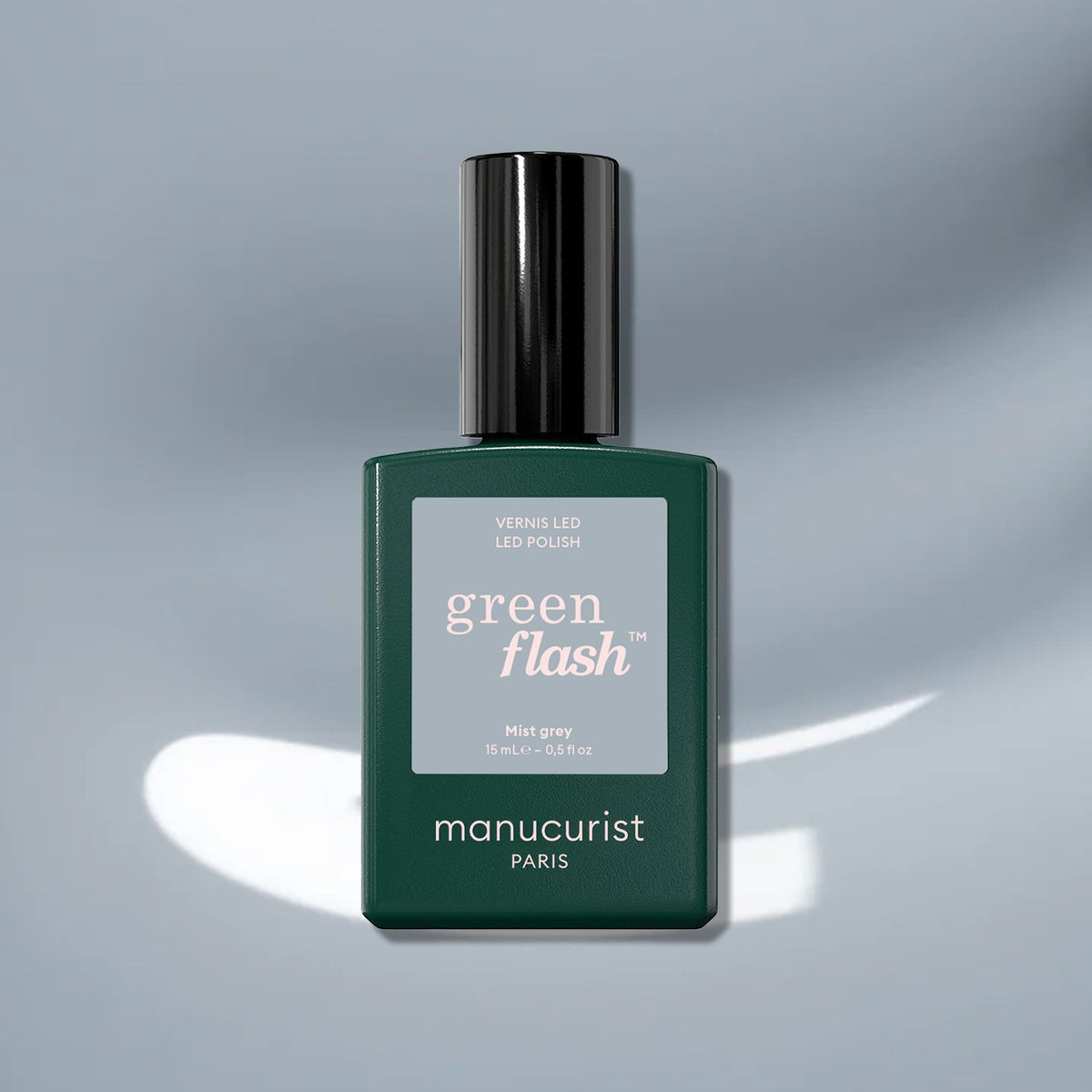 A bottle of Manucurist Mist Grey sits on a shiny light grey surface with white reflections, its dark cap and teal green label highlighted by muted blue tones that add elegance to the scene.