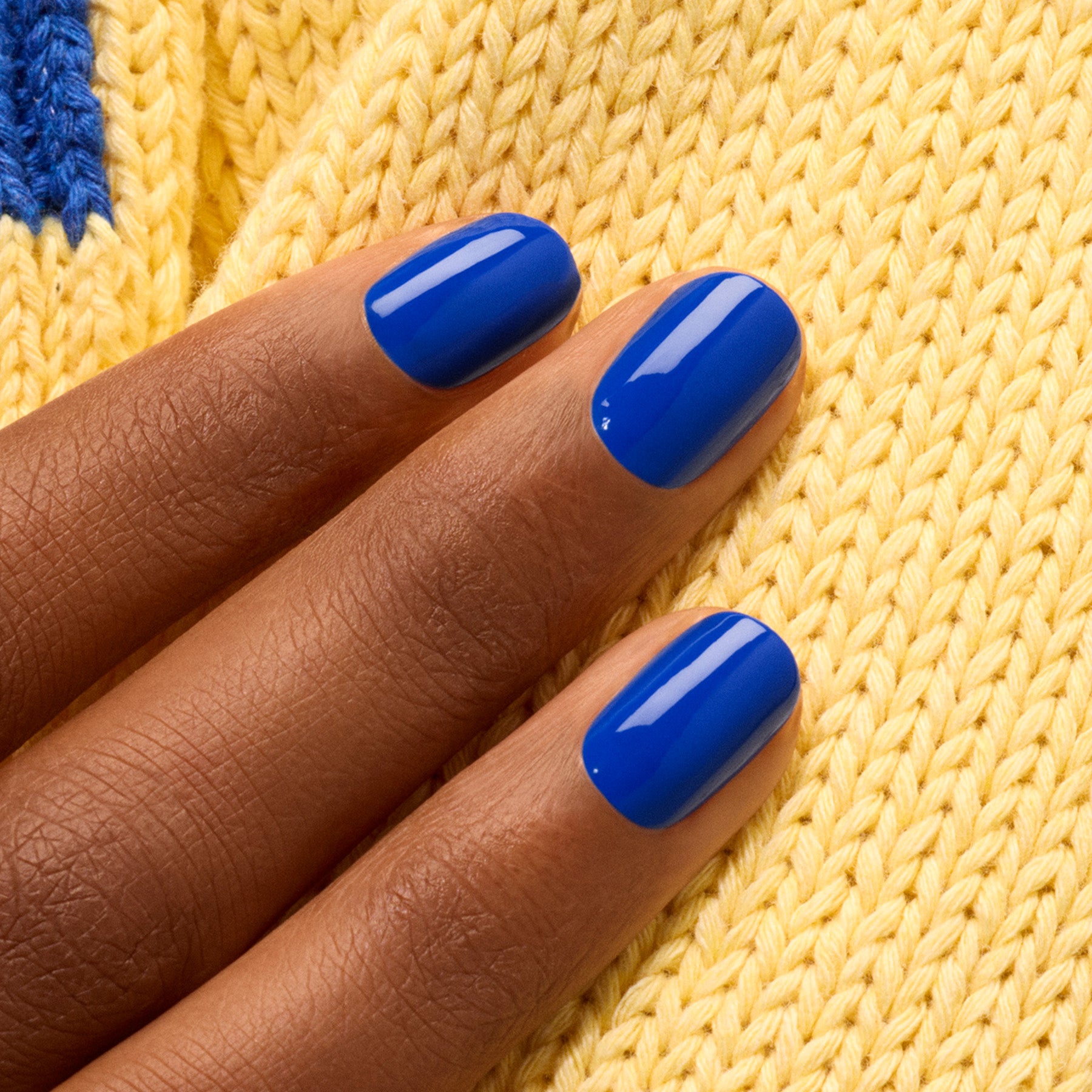 A hand with neatly manicured nails painted in Manucurist’s Ultramarine polish rests on yellow knitted fabric.
