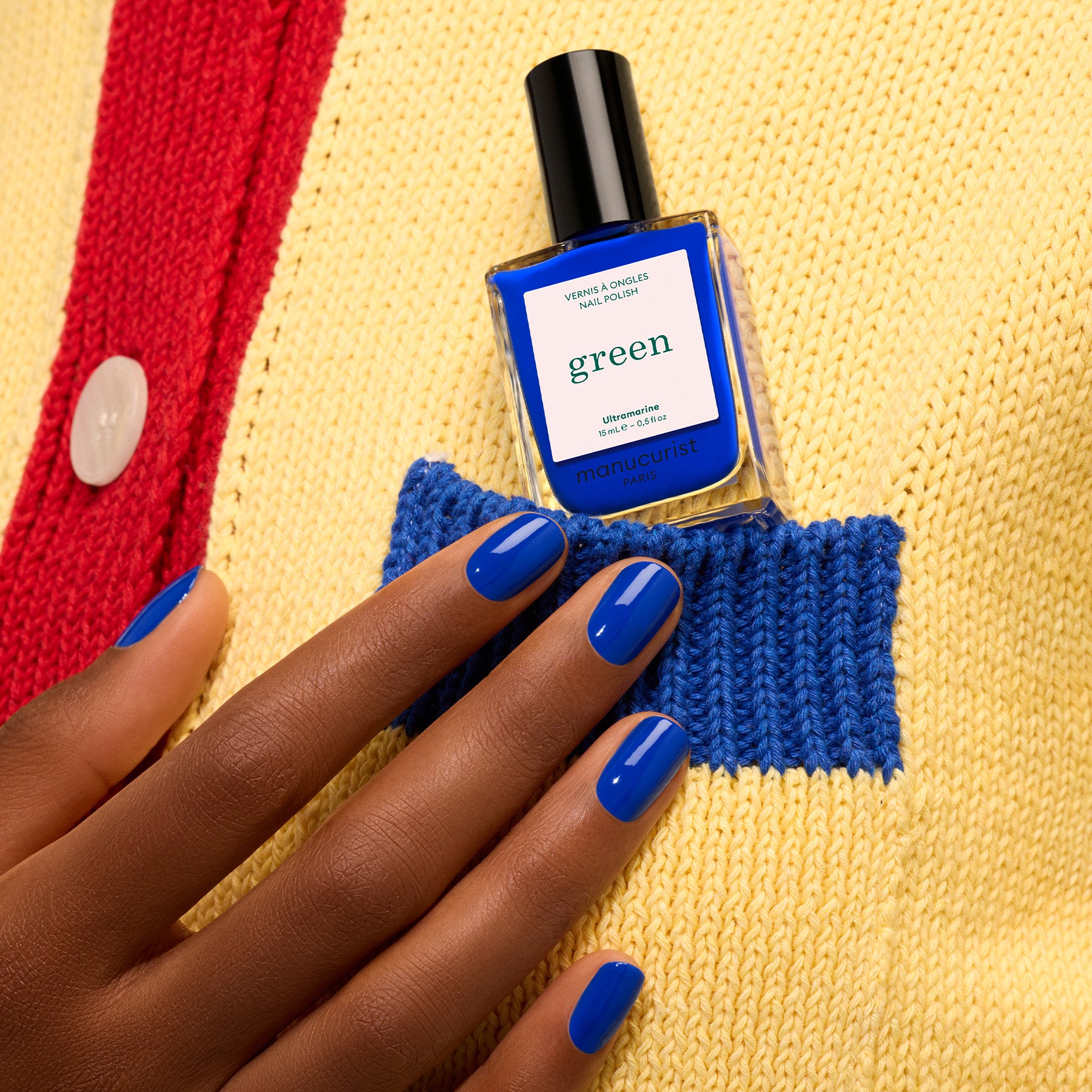 A hand with Ultramarine ocean blue nail polish holds a matching Manucurist bottle from the Green™ range, tucked into the pocket of a yellow and blue knit sweater with a red stripe and white button.