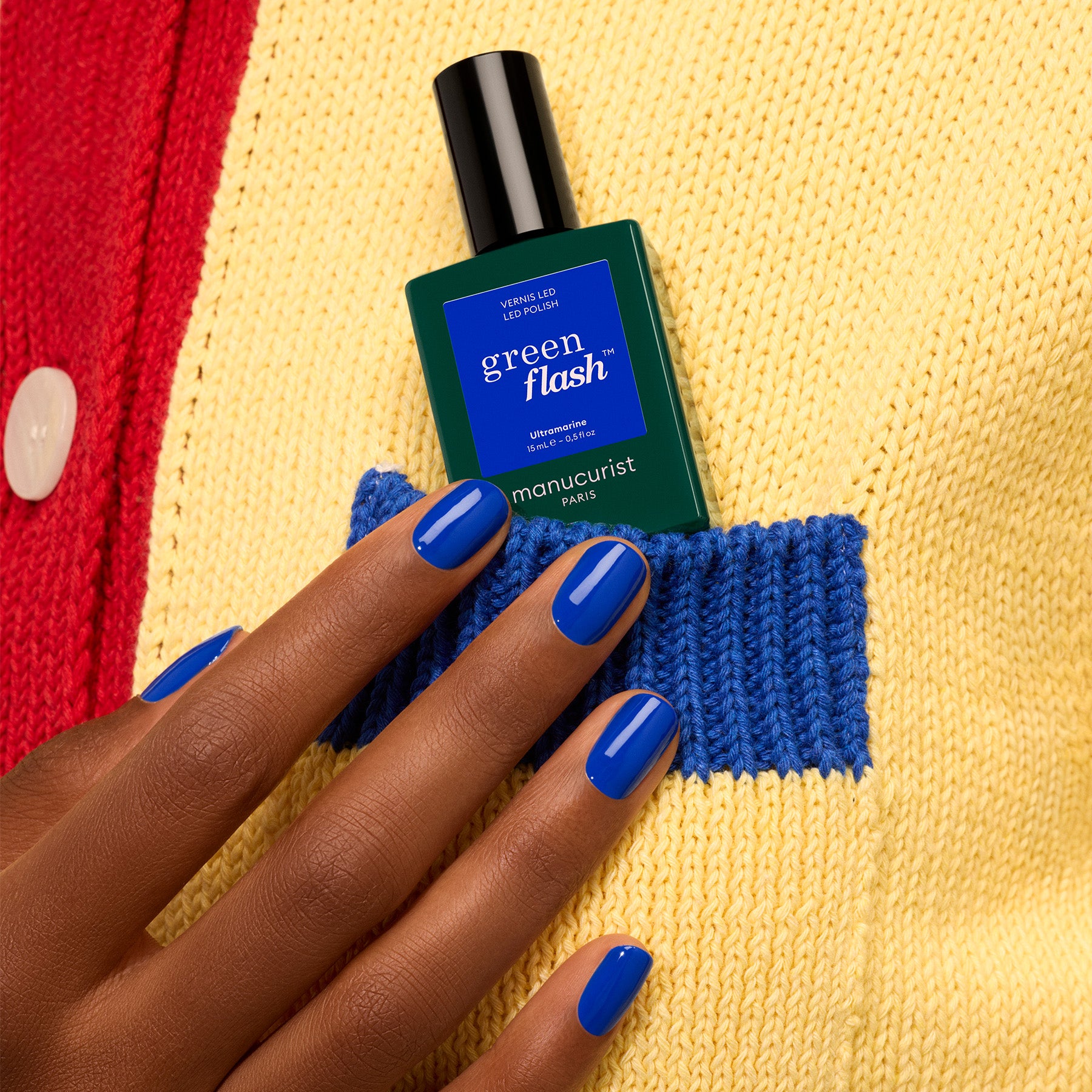 A hand with glossy electric blue gel polish (Ultramarine by Manucurist) holds a green bottle of Green Flash nail polish near the blue pocket of a yellow and red knitted sweater.