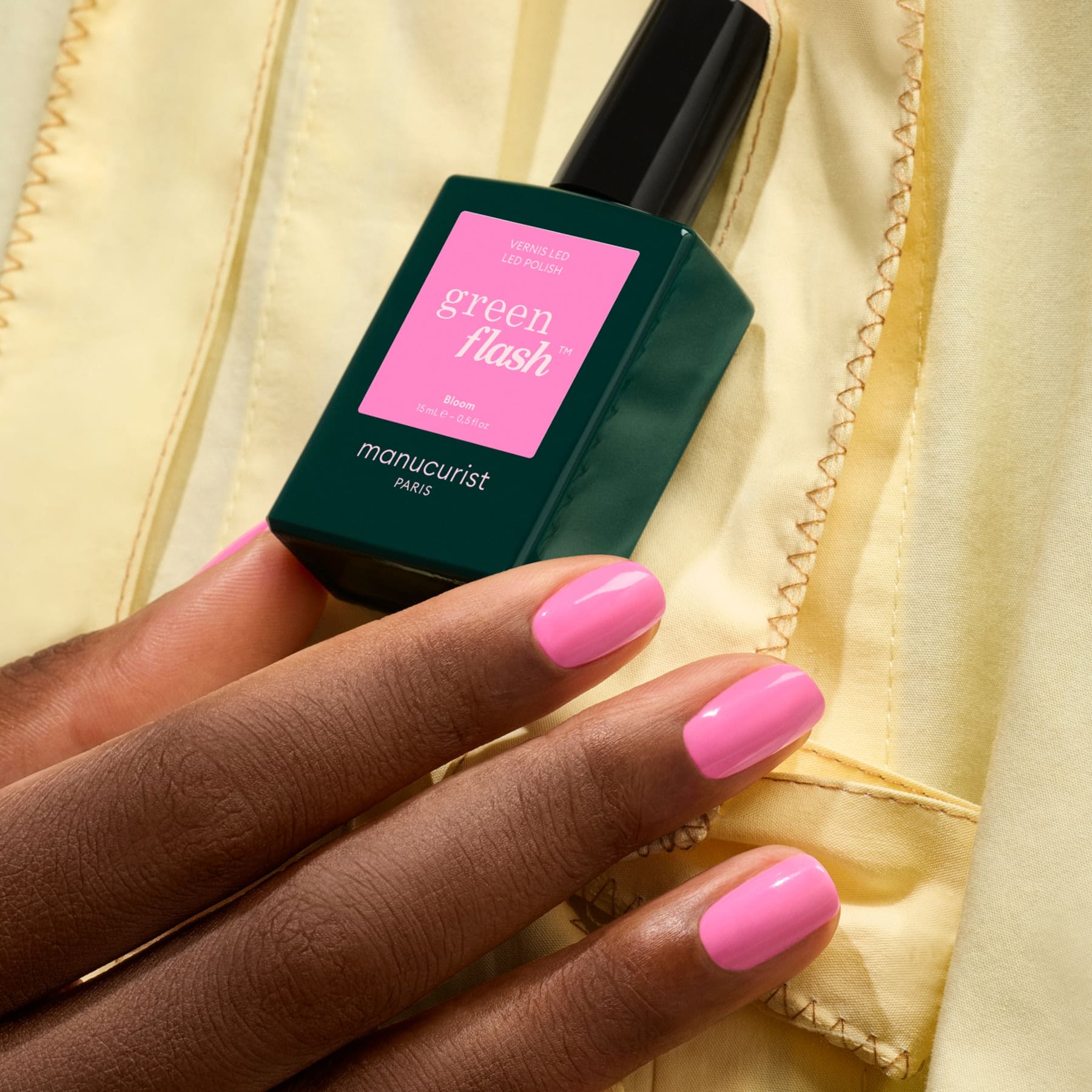 A hand with neatly manicured nails painted neon pink holds a green bottle of Manucurist Bloom gel polish against a yellow fabric background.