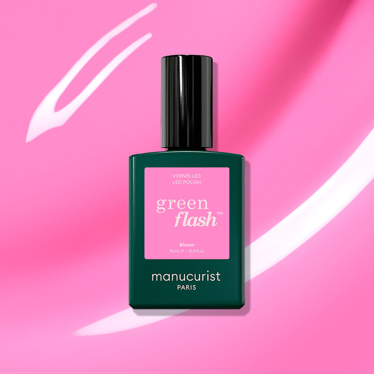 A bottle of Manucurist Bloom gel polish sits against a vibrant pink background with white streaks. The dark green bottle and pink label highlight the chic, magnetic intensity of this nail color.