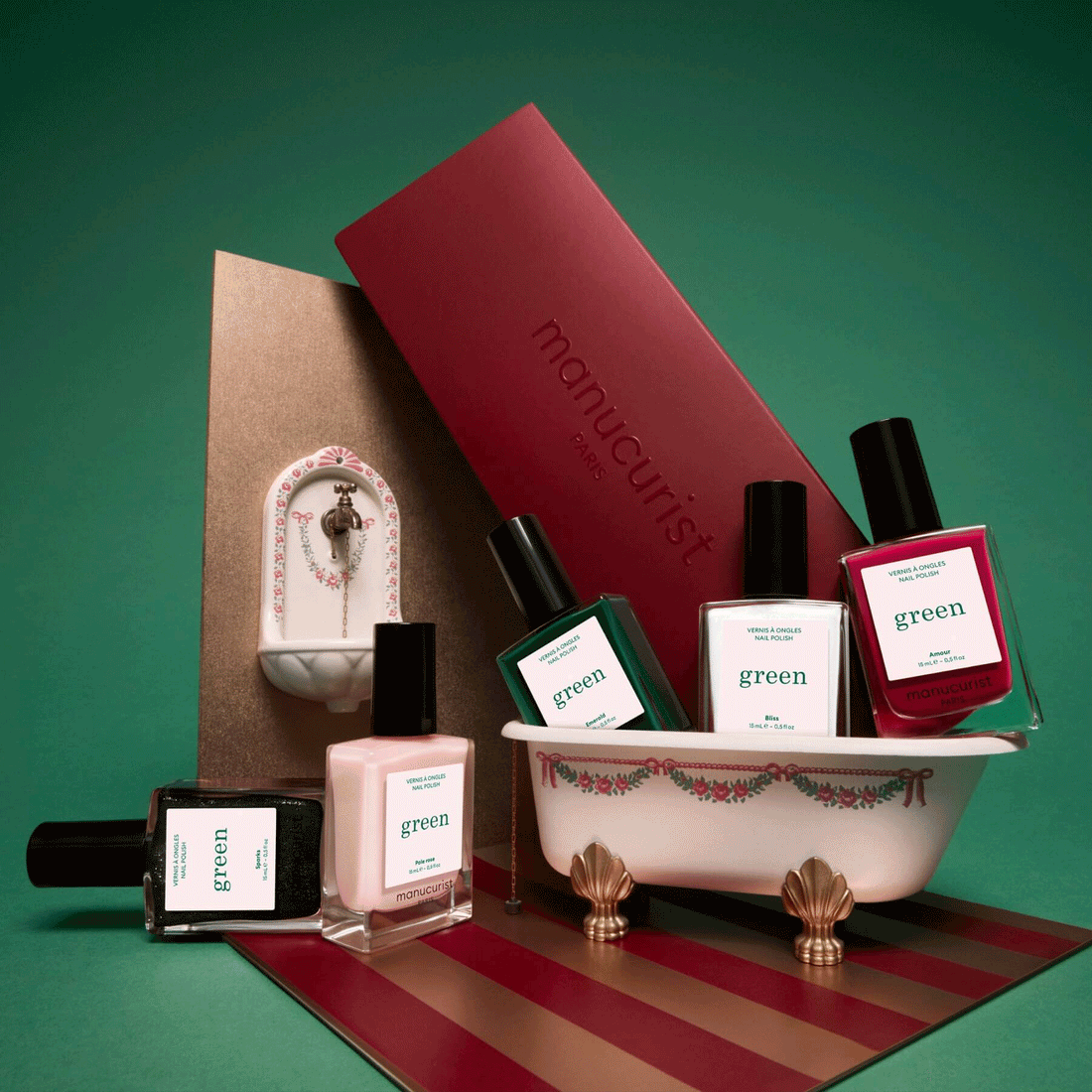 Five bottles of Manucurist’s Customisable Rainbow Green nail polish are arranged around a small decorative bathtub, with a red manicure kit and a miniature sink in the background, all set against a green backdrop—creating the perfect display for any nail polish collection.