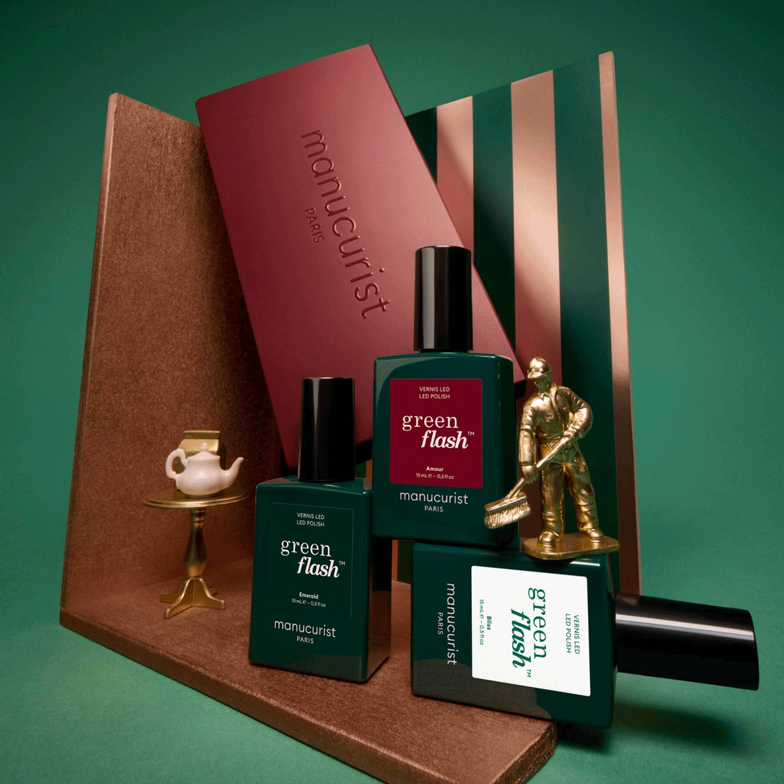 Three green bottles labeled Customisable Trio Green Flash by Manucurist are arranged with a gold figure, a small tea set, and striped and maroon props on a green background—perfect inspiration to create your own trio with their acetone-free gel polish remover for a complete manicure experience.