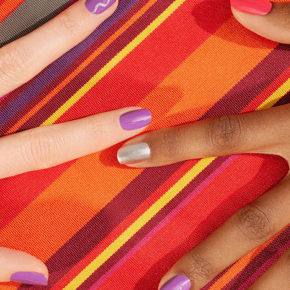 Two hands with manicured nails in Manucurist’s Violette shade rest on vibrant striped orange and red fabric, silver and coral accents highlighting the smooth, lasting shine of Green Flash gel polish.