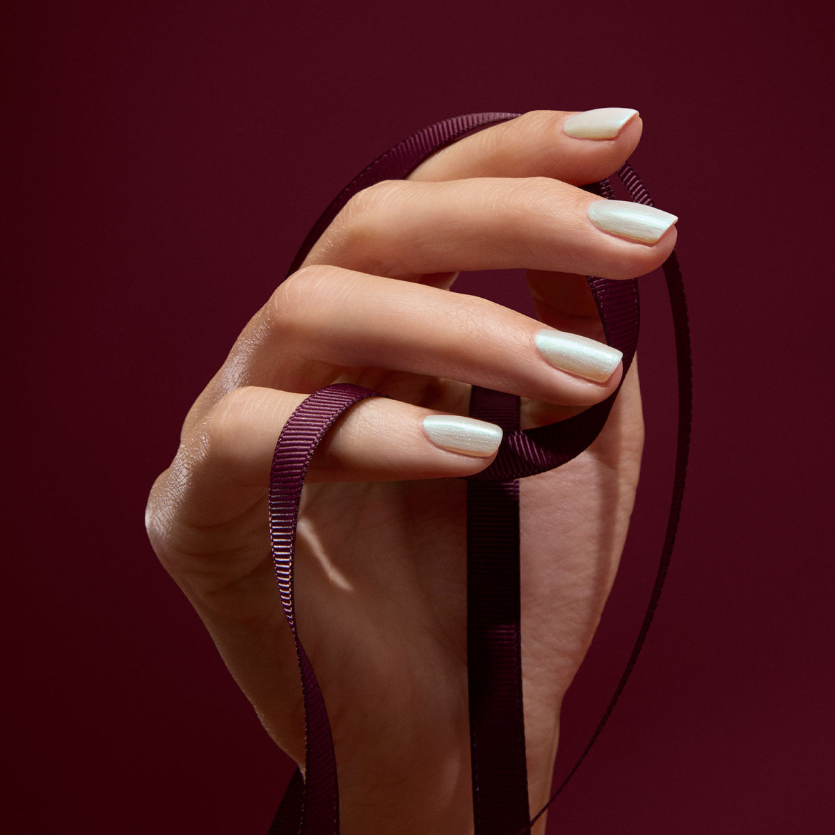 A hand with Fairy pearly white gel polish by Manucurist holds a dark burgundy ribbon against a matching background.