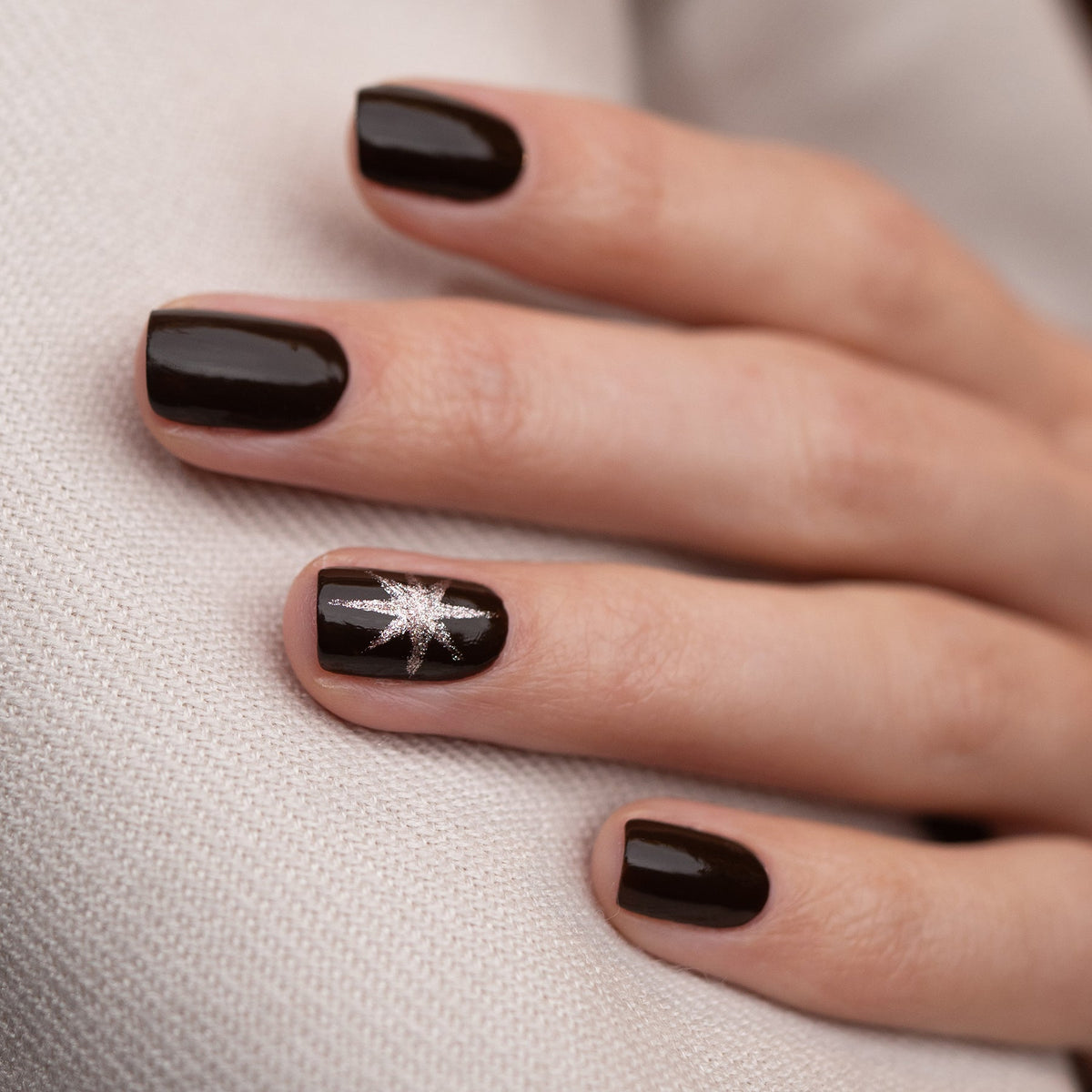 A hand with short, neat nails painted in Manucurist’s Clove—a rich chocolate brown polish—rests on light textured fabric. The ring finger features a silver starburst design.