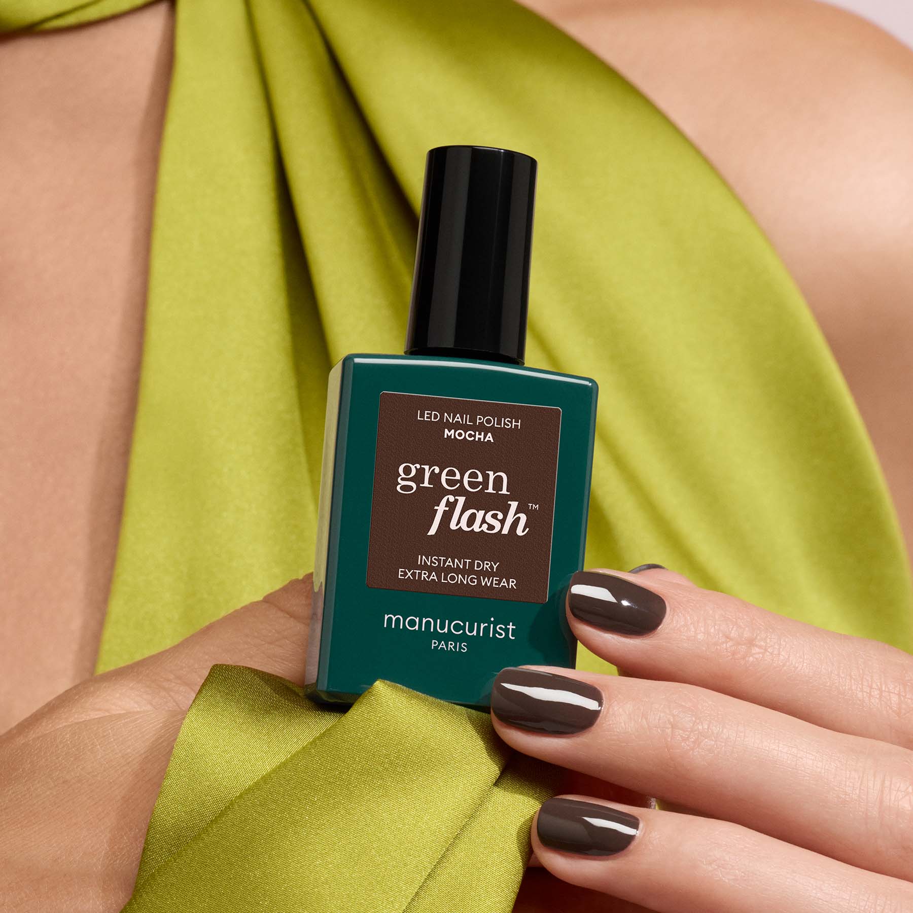 A person with manicured brown nails holds a bottle of Manucurist Mocha nail polish, a dark brown shade. They wear lime green, silky fabric draped around their neck and shoulders.