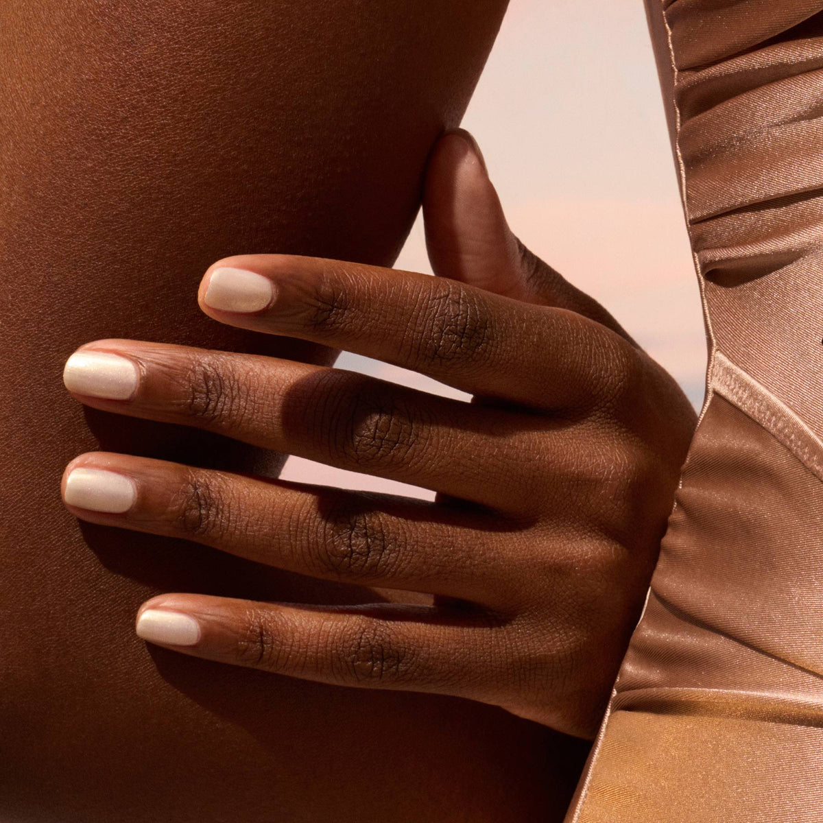 A close-up of a hand with neatly manicured nails polished in Manucurist’s Solar shade rests gently on a bare thigh. The person wears a silky, light brown garment under soft, warm lighting.